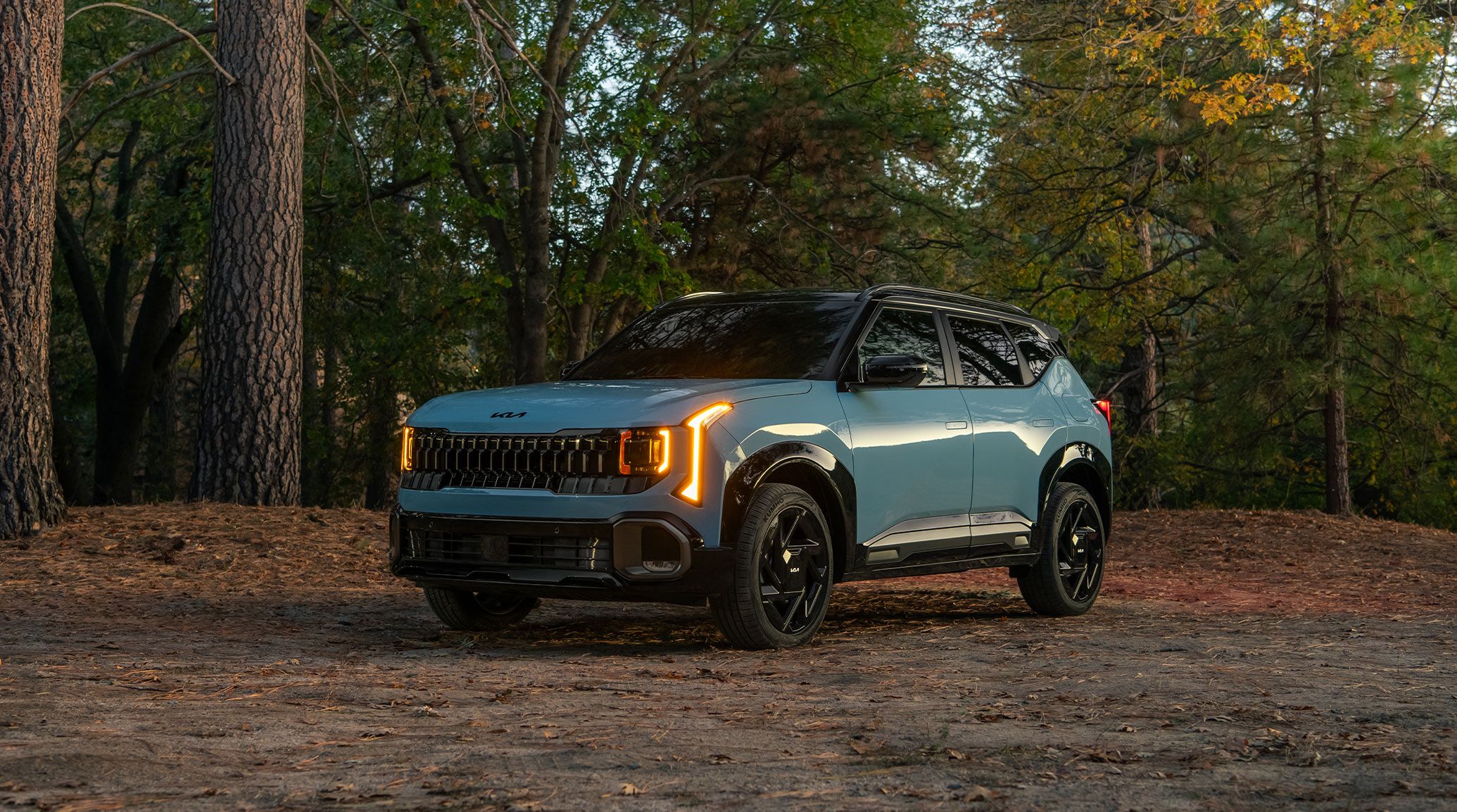 Side view of the 2027 Kia Seltos compact SUV parked in a forest with lush trees behind the vehicle. This photograph shows the gloss black exterior trim, as well as the bright orange LED headlights and roof rails.