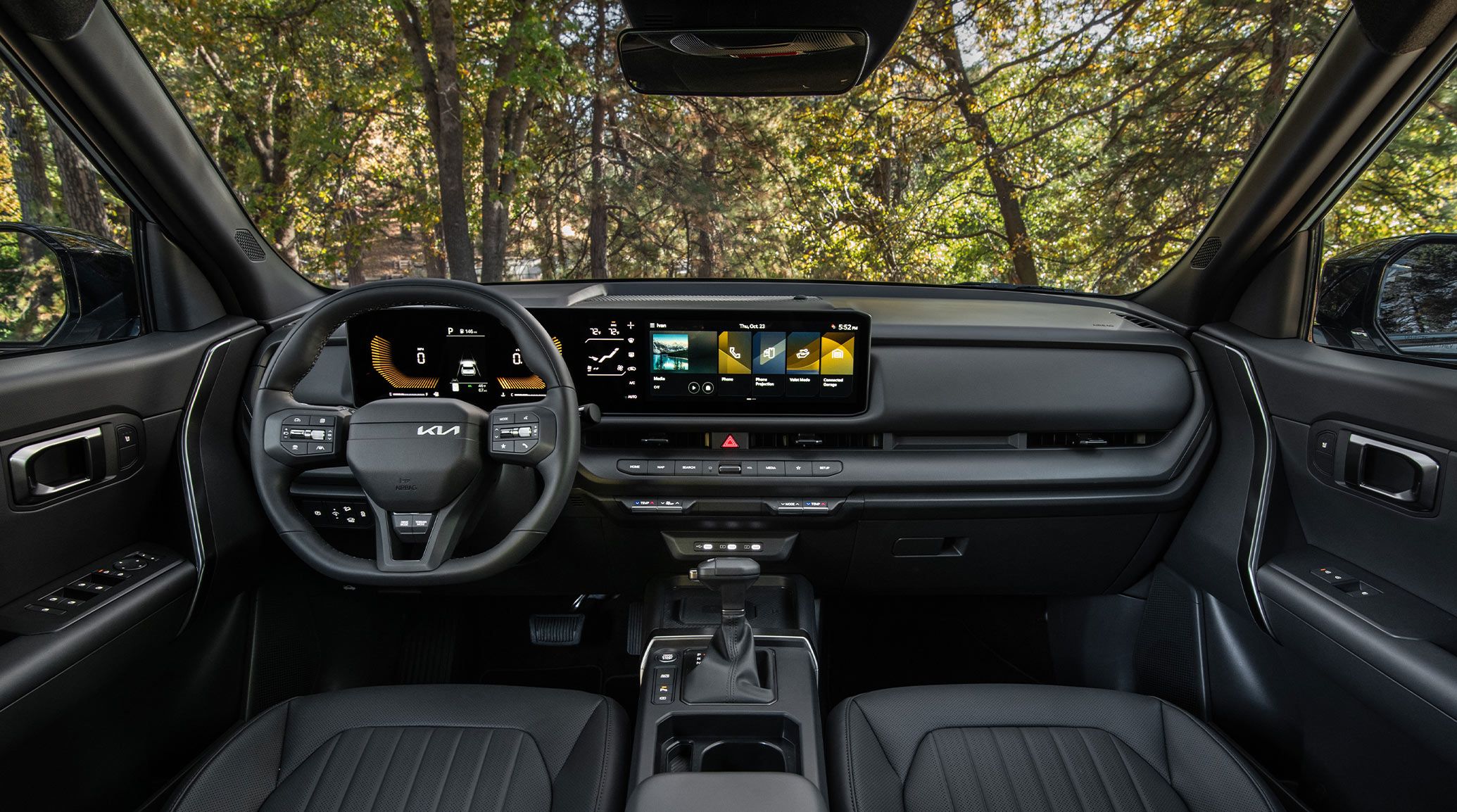 Interior wide-angle photo inside the 2027 Kia Seltos showcasing its steering wheel, dashboard display, center console, shifter, rear view mirrors, glove compartment, and more. A forest is visible through the windshield.