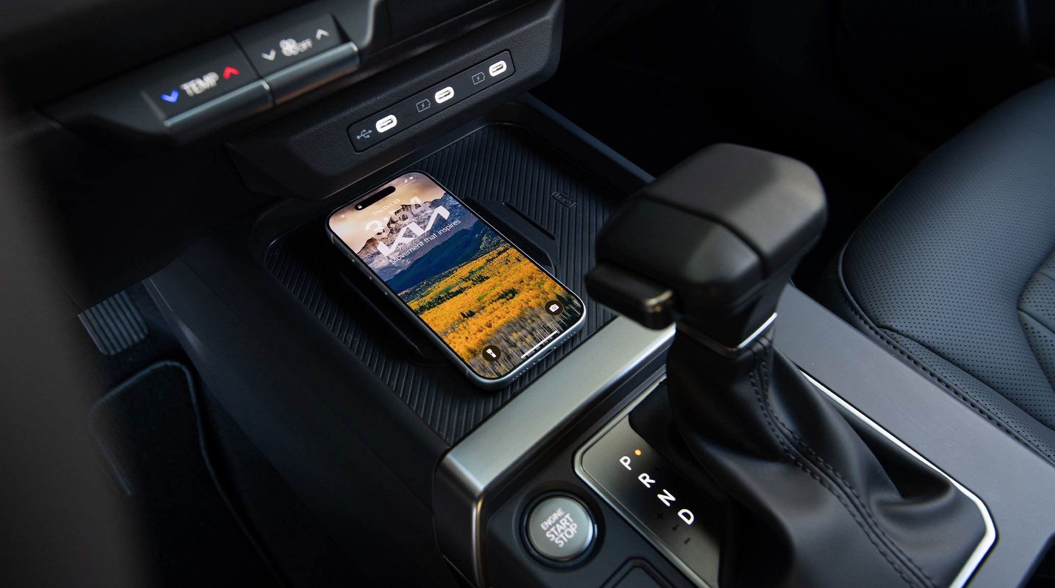 Close-up interior view of the 2027 Kia Seltos gear shifter in park and wireless charging station holding a charged smartphone with the Kia logo and mountains as the lock screen, highlighting the modern tech features.
