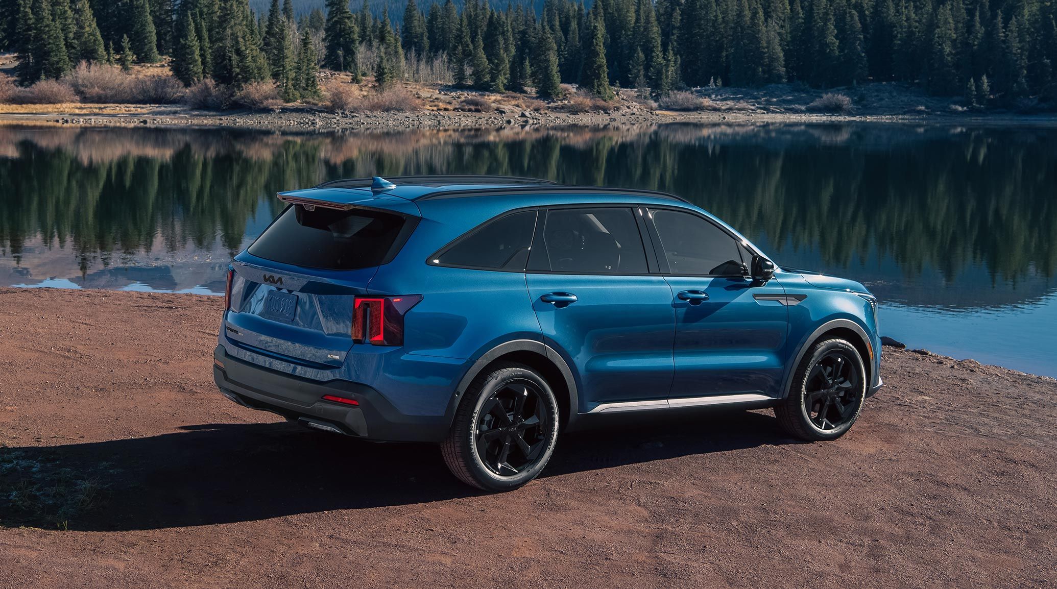 Rear-passenger-side view of the 2027 Kia Sorento Hybrid SUV in blue, shining sleekly in the sunlight, parked on a dirt surface near a pond reflecting wilderness in the background.