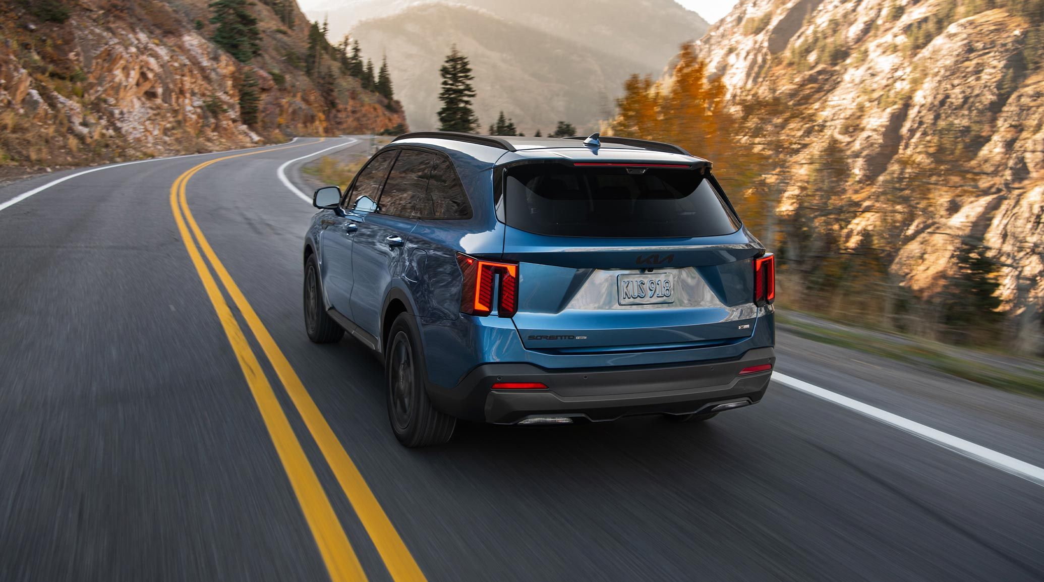 Rear driver-side view of the 2027 Kia Sorento Hybrid SUV in blue as it drives around a curving mountain road, with rocks and trees visible in the background. The sleek angles and lines of the Sorento Hybrid are plainly visible.
