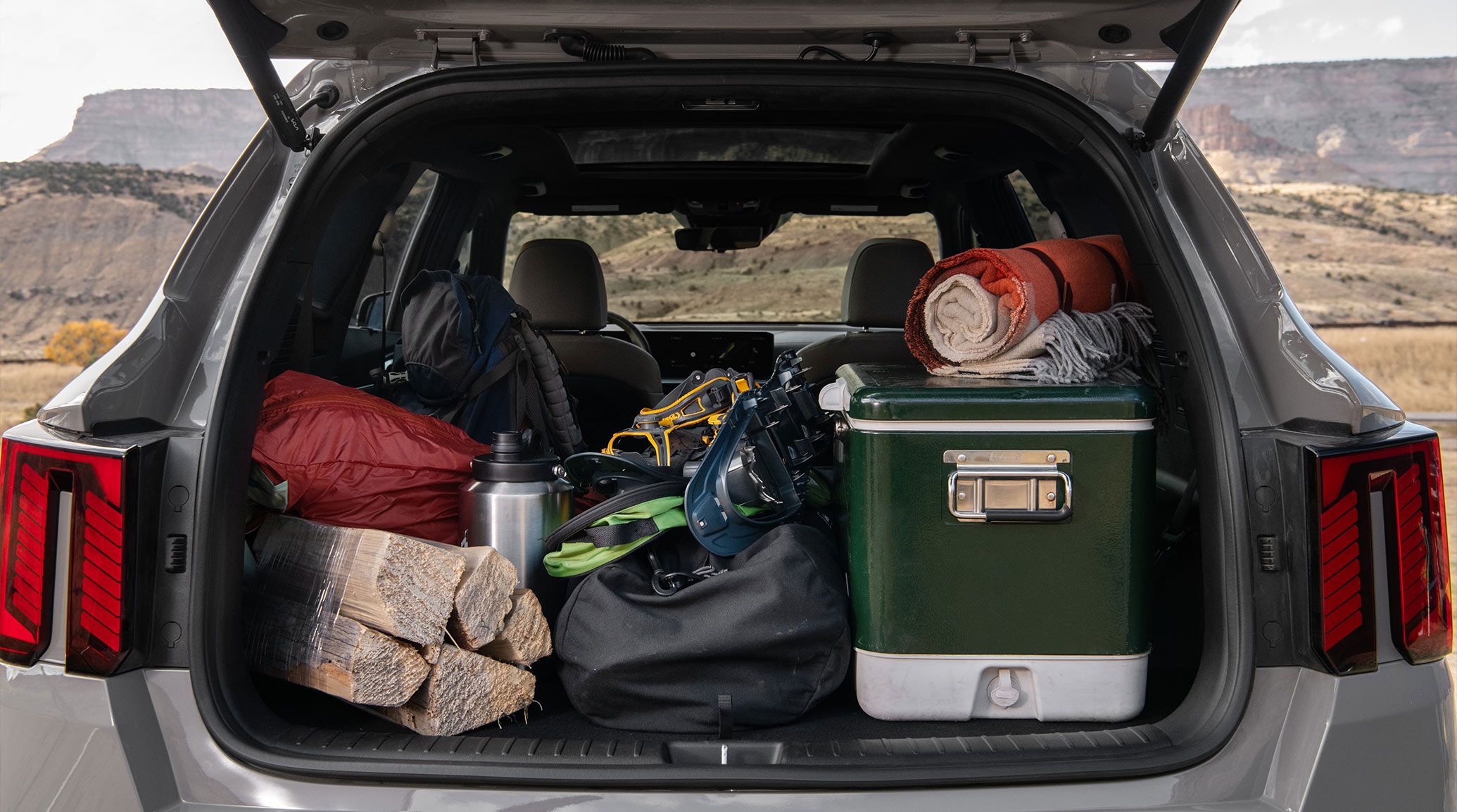 Close-up photo of the 2027 Kia Sorento Hybrid SUV with an open trunk showcasing the spacious cargo area packed with outdoor gear for camping, set against a hillside backdrop.