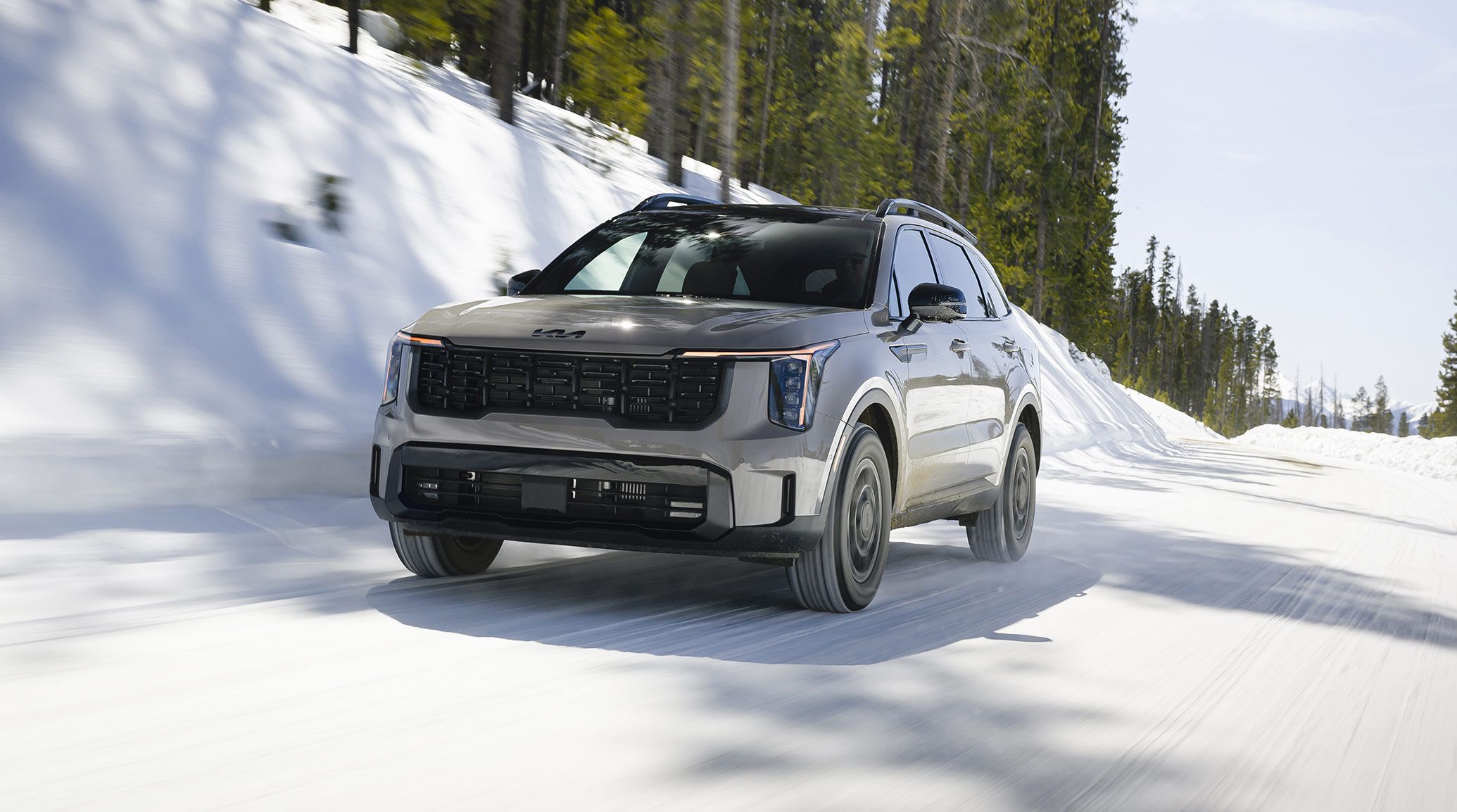 Angled shot of the 2027 Kia Sorento in gray, highlighting the black accents, all-wheel drive, and sleek design while cruising through a snow-covered road with trees behind it.