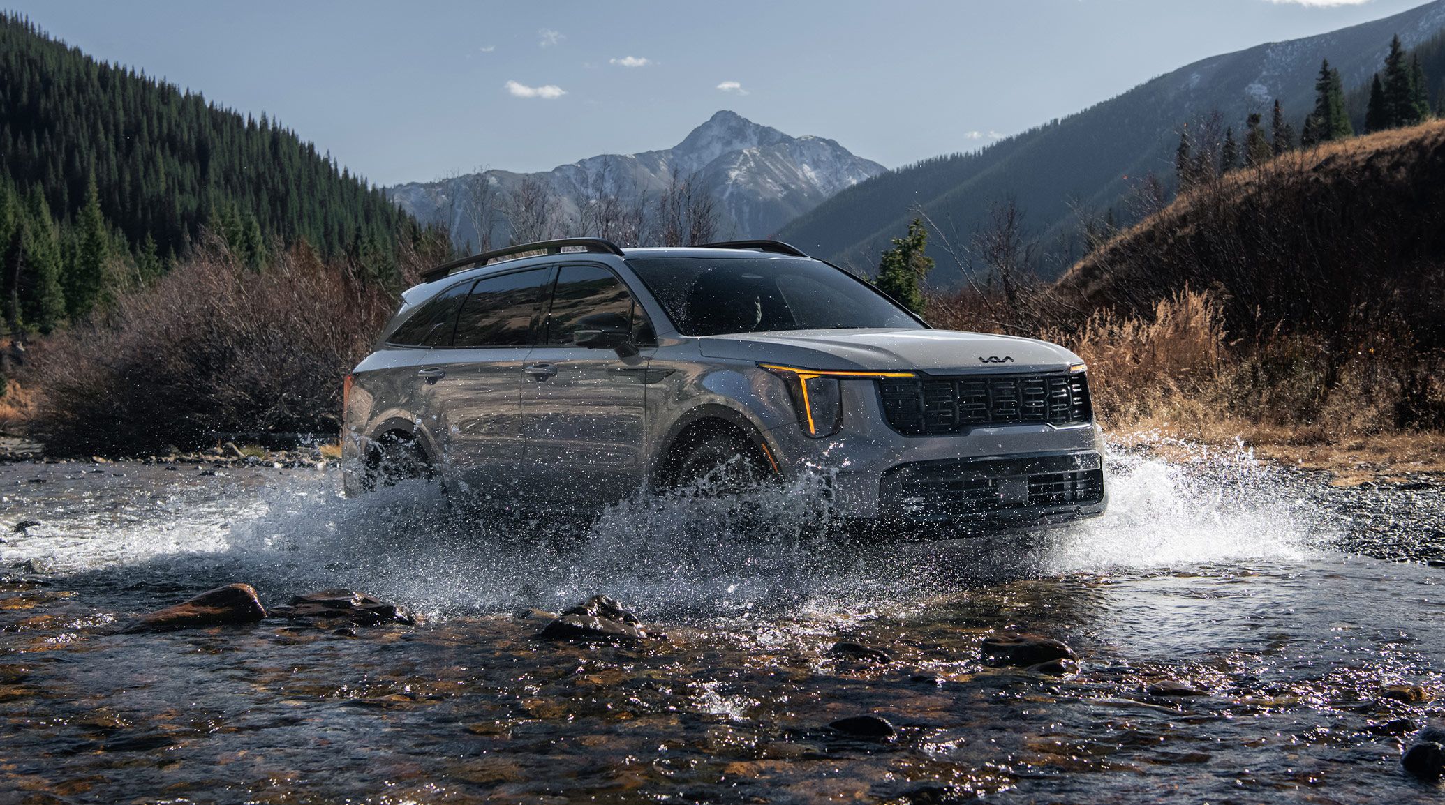 Action shot of a gray 2027 Kia Sorento in a three-quarter view with mountains and greenery in the backdrop. The SUV is driving through a river and is making a big splash, highlighting the all-terrain tires and unique front fascia.