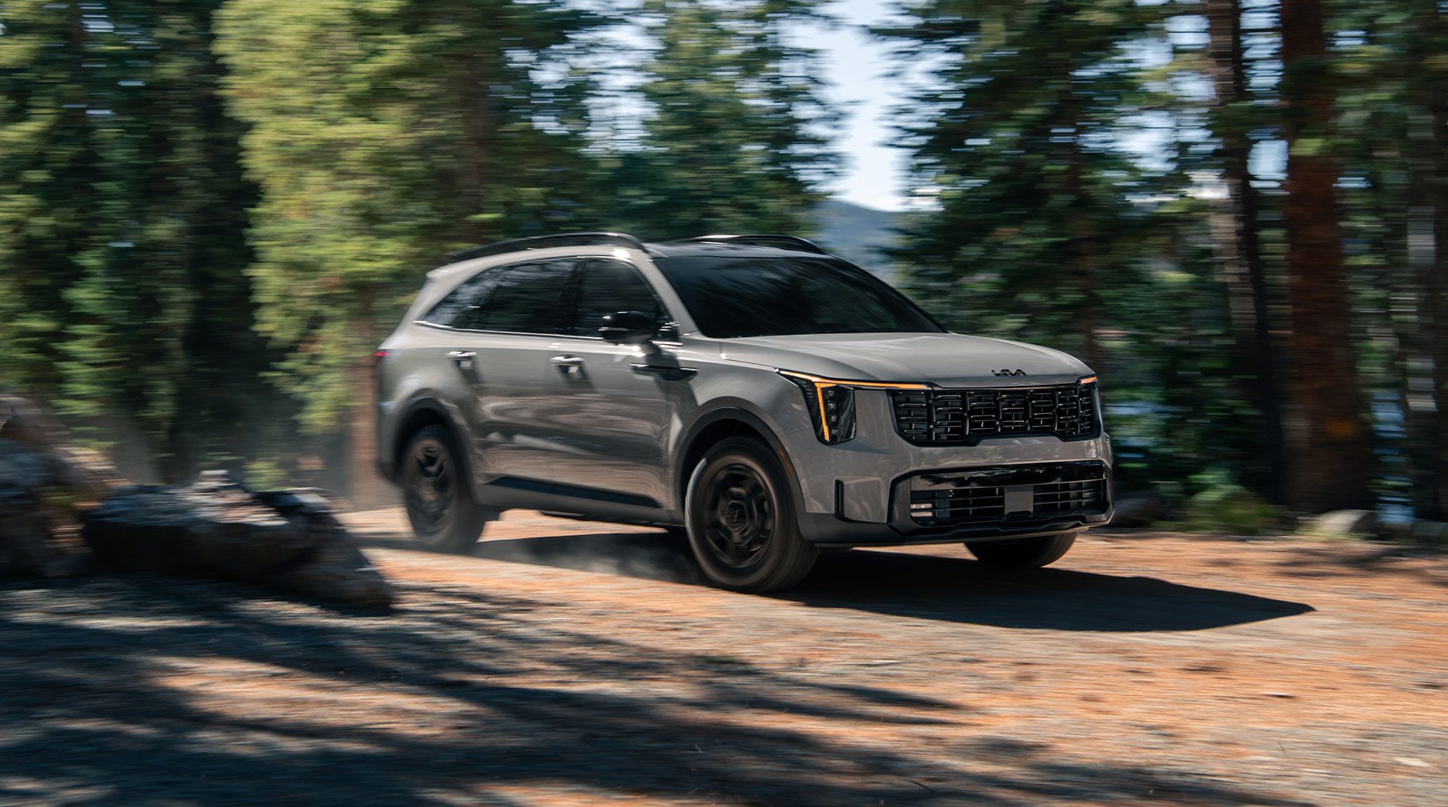 Dynamic shot of the 2027 Kia Sorento in gray speeding along a forest road with trees and rocks in the background, featuring a bold front grille, striking LED headlights, and the available turbocharged engine.