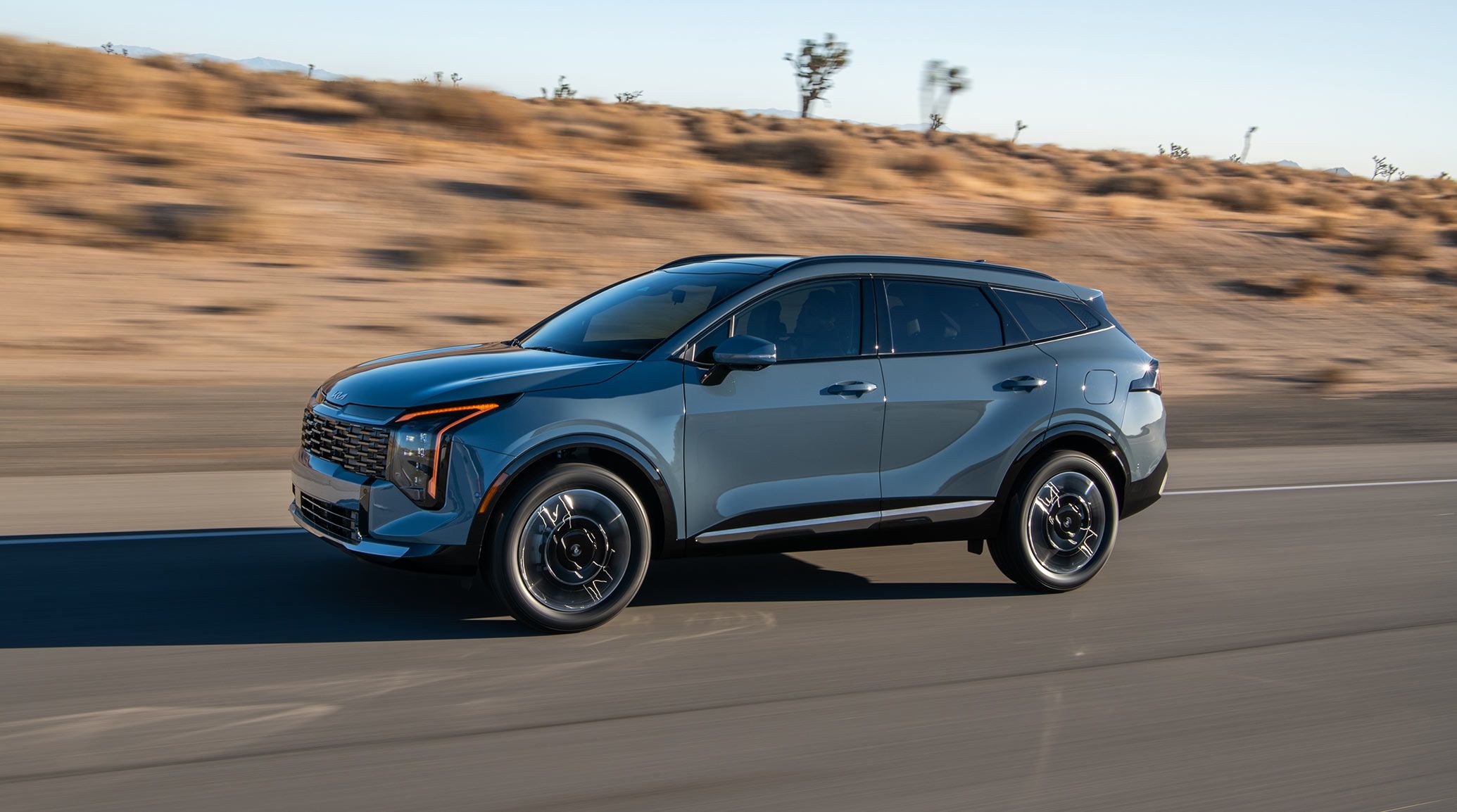 A blue 2027 Kia Sportage Hybrid compact SUV is driving along a desert highway, highlighting the driver’s side and sleek design.