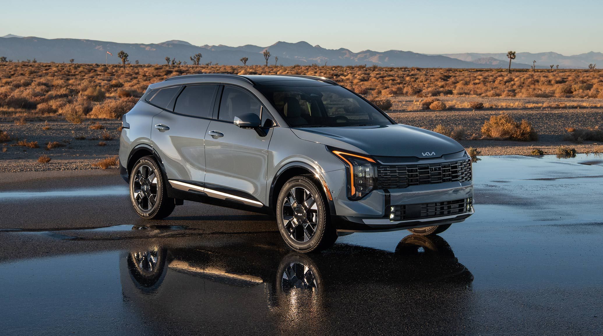 Front three-quarter view of the 2027 Kia Sportage Hybrid SX Prestige in blue parked on a wet road with desert landscapes and mountains in the background. The shot is highlighting the compact SUV’s vertical LED headlights and amber daytime running lights.