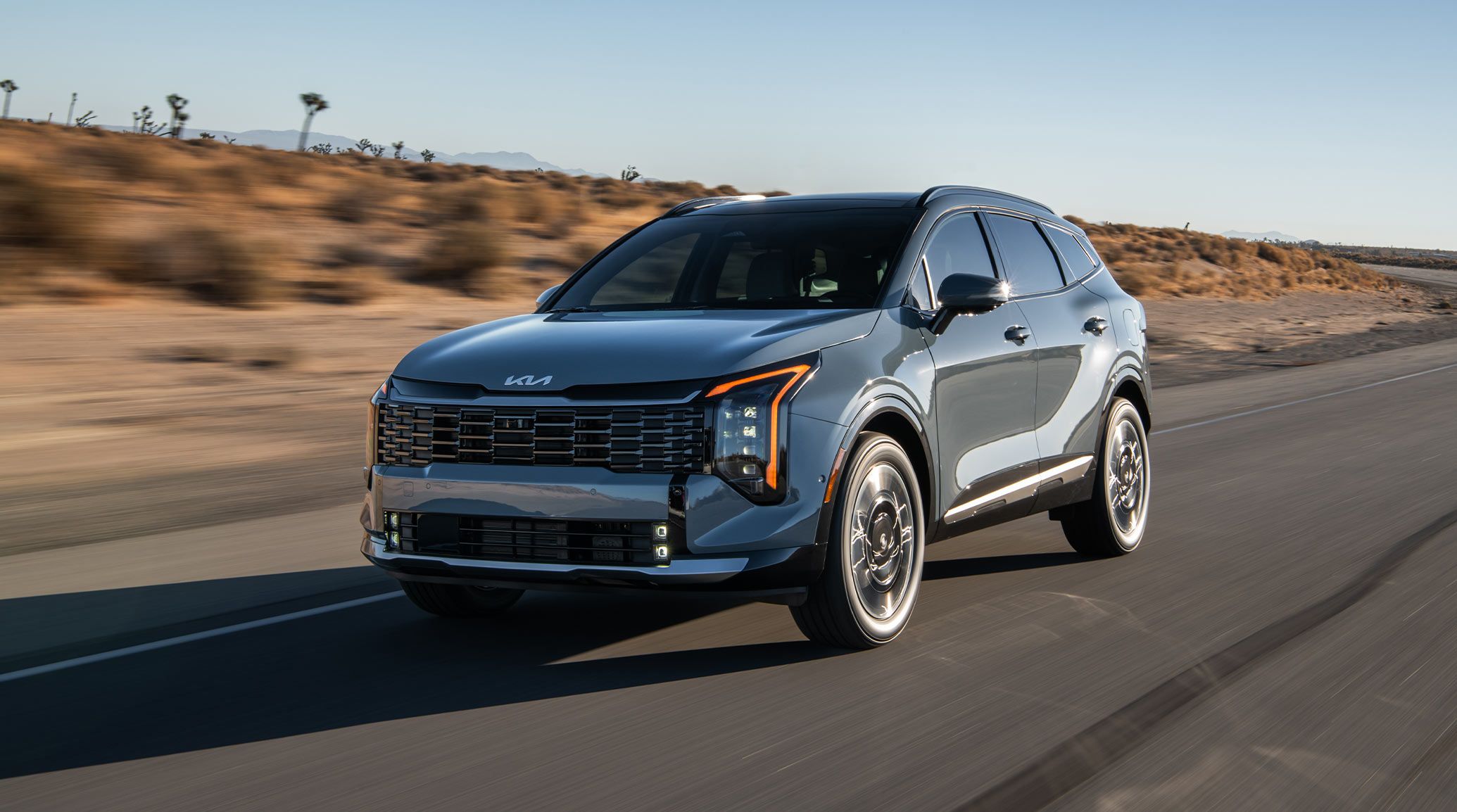 2027 Kia Sportage Hybrid driving on a desert highway, shown in a front three‑quarter view with vertical LED headlights, sculpted body lines, and distinctive wheel styling.