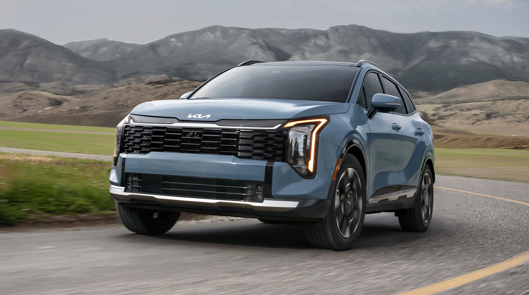 Dynamic shot of the 2027 Sportage Hybrid in blue speeding along a curvy scenic mountain road, featuring a bold front grille, striking LED headlights, and distinctive wheel designs.