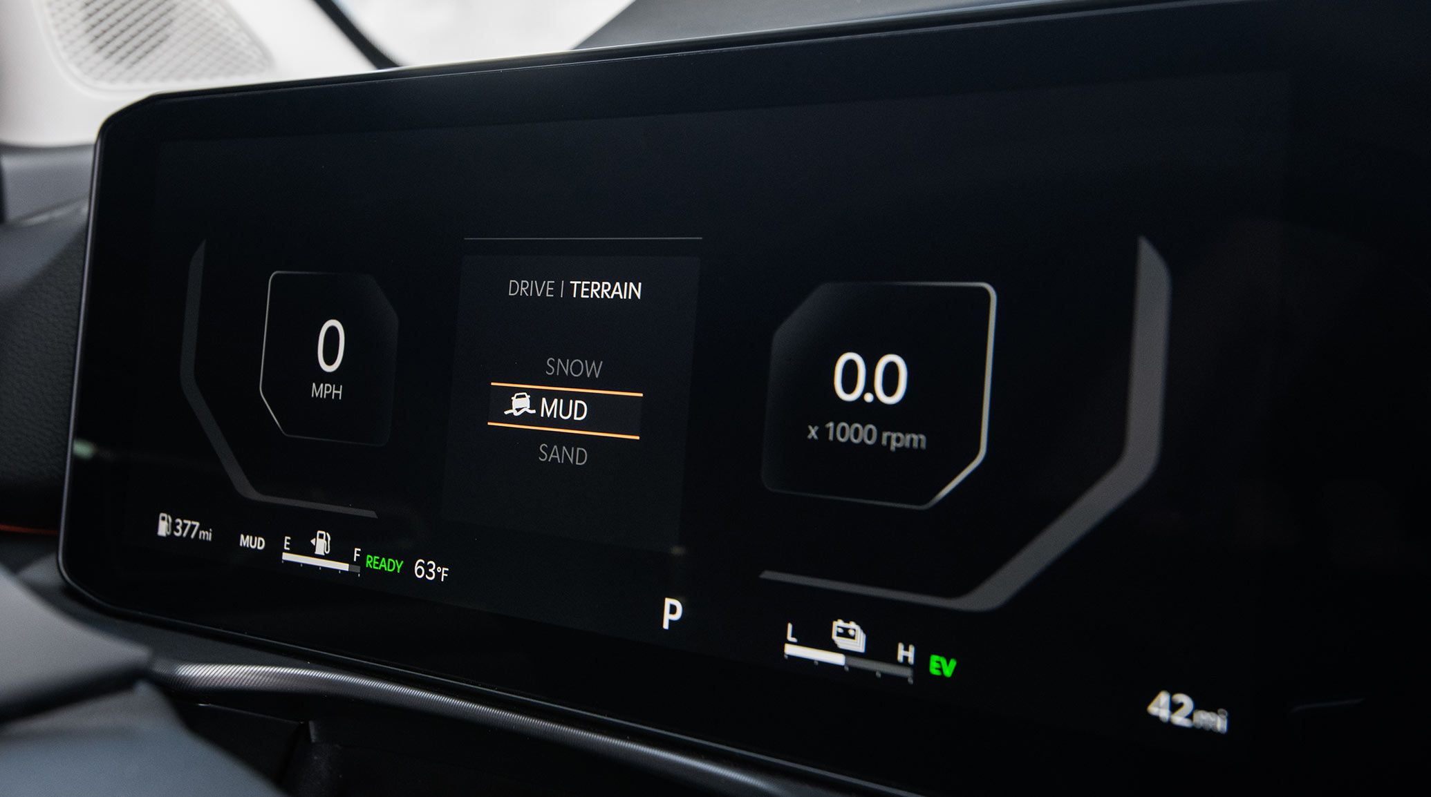 Close-up of the 2027 Kia Sportage Hybrid interior dashboard displaying miles per hour on the left, terrain mode in the middle with mud selected, and RPM on the right, highlighting the available all-wheel drive.
