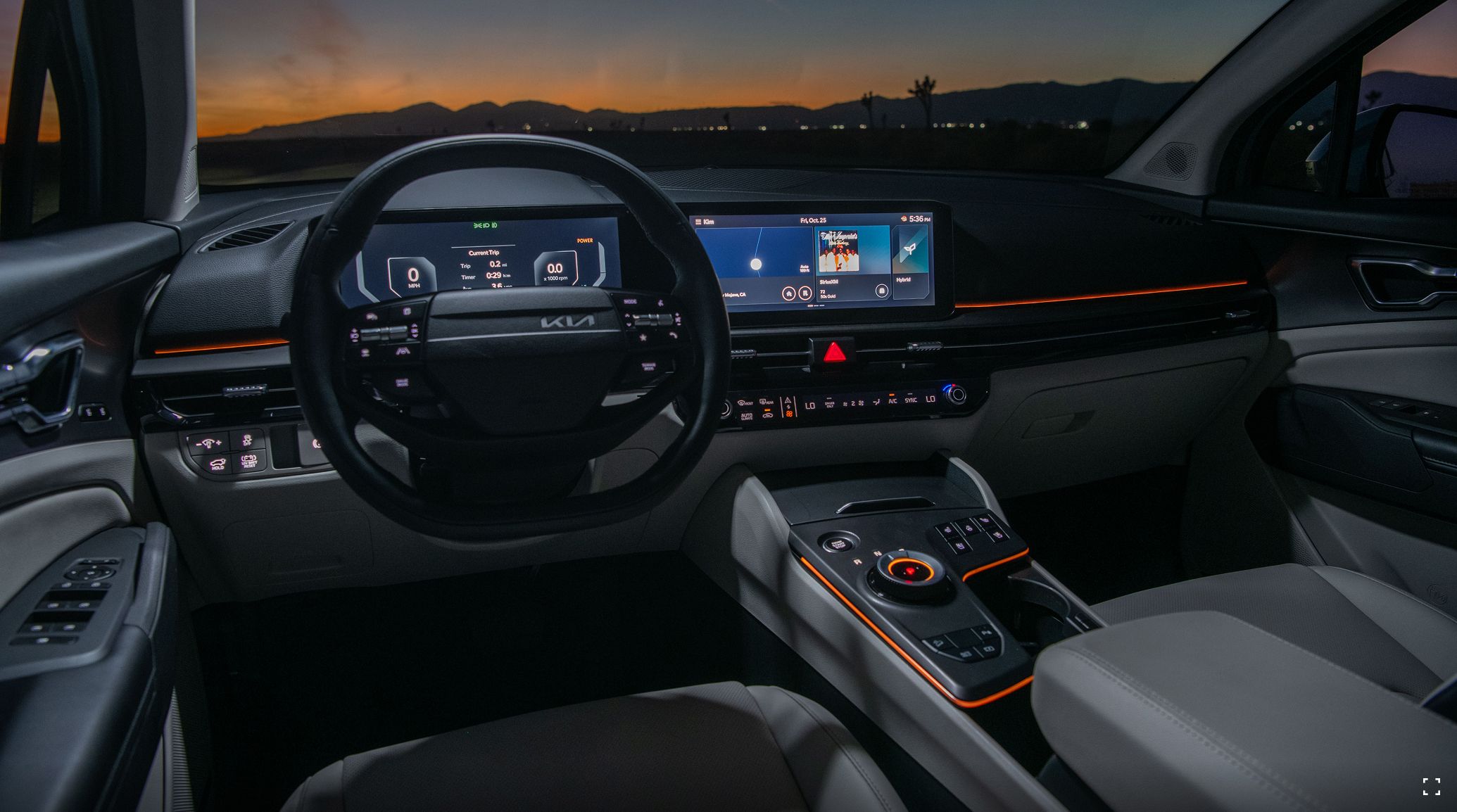 Interior view of the 2027 Kia Sportage Hybrid cockpit with a digital instrument cluster, wide infotainment display, and color‑selectable ambient mood lighting set to red, illuminating the dashboard and center console with a sunset and mountains visible through the windshield.