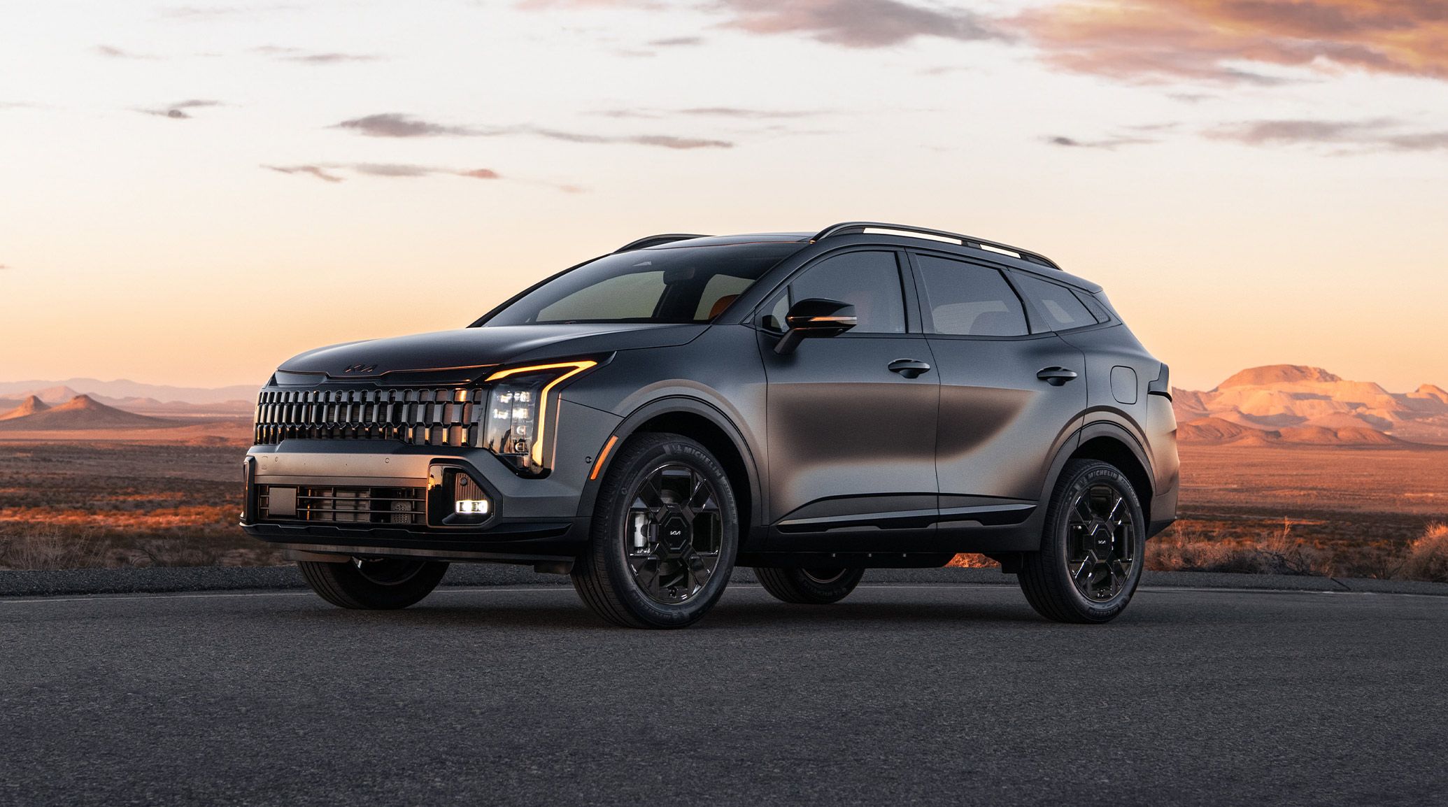 A still shot of the 2027 Kia Sportage Plug-In Hybrid in gray parked on a road during sunset with towering mountains in the background, highlighting the driver’s side with bold headlights, alloy wheels, and high-utility roof rails.