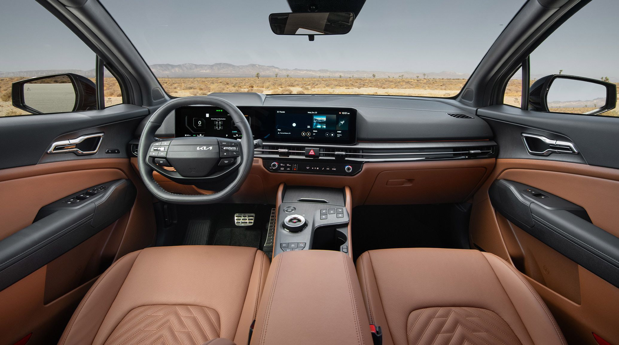 Interior view of the 2027 Kia Sportage Plug-In Hybrid X-Line Prestige in brown featuring the steering wheel, digital display with maps and music showing, and the center console with a scenic desert landscape and mountains visible through the windshield.