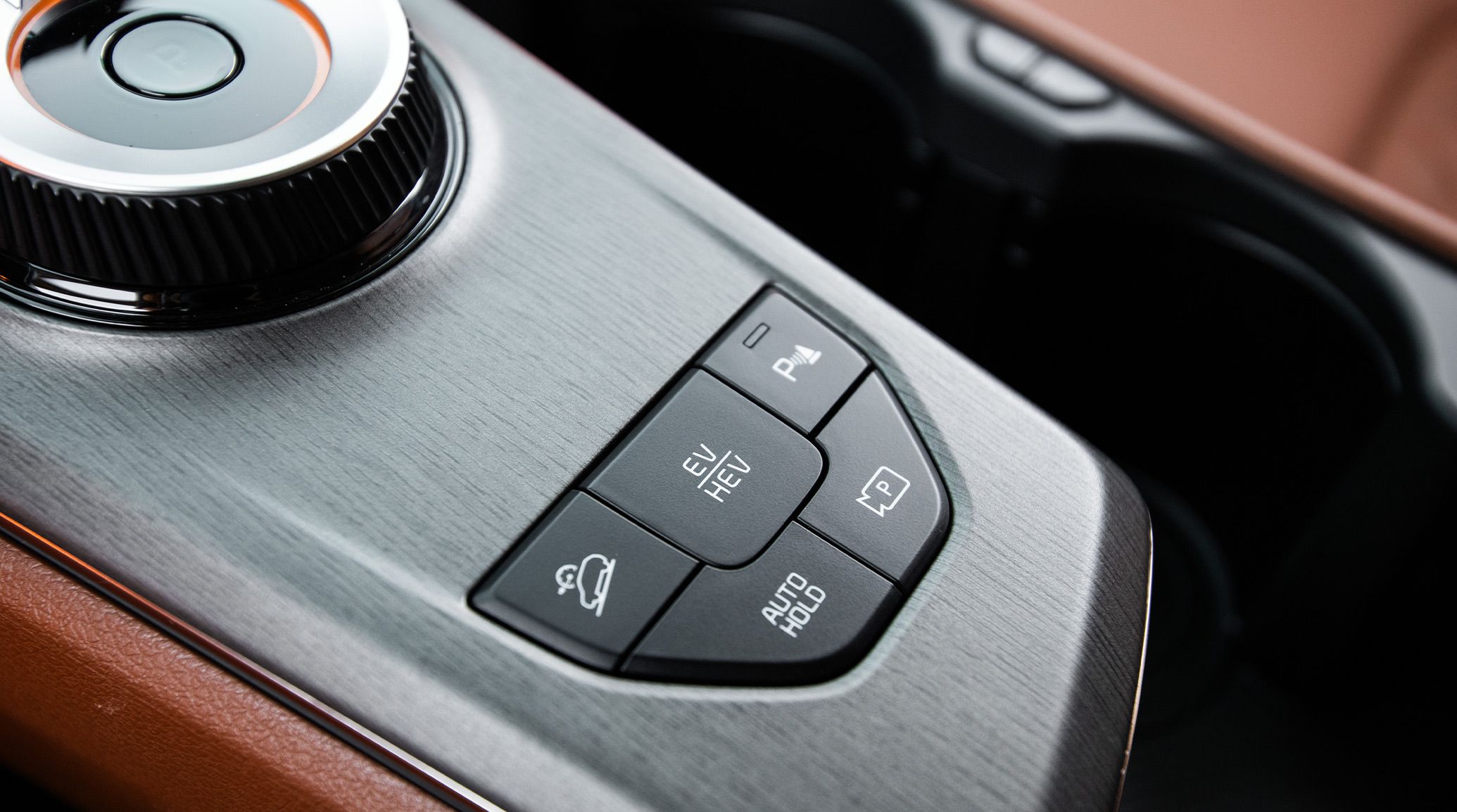 Close-up of the silver rotary shifter dial in the 2027 Kia Sportage Plug-In Hybrid. The image also features 5 buttons that include electric mode, hybrid mode, and more. Cup holders and the brown interior are visible in the background.