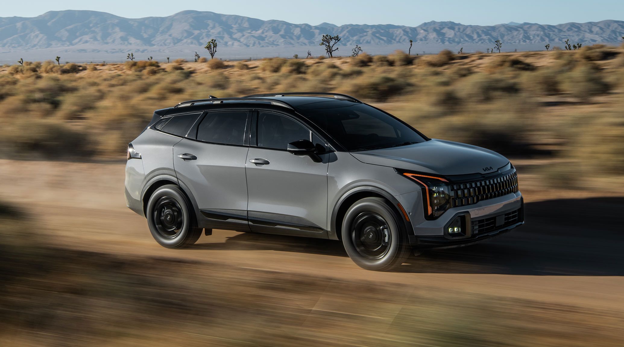 An exciting action shot of the 2027 Kia Sportage SUV speeding over desert scrubland. The vehicle's curves, angles, roof rack, and grille are especially prominent. Mountains and Joshua trees are visible in the background.