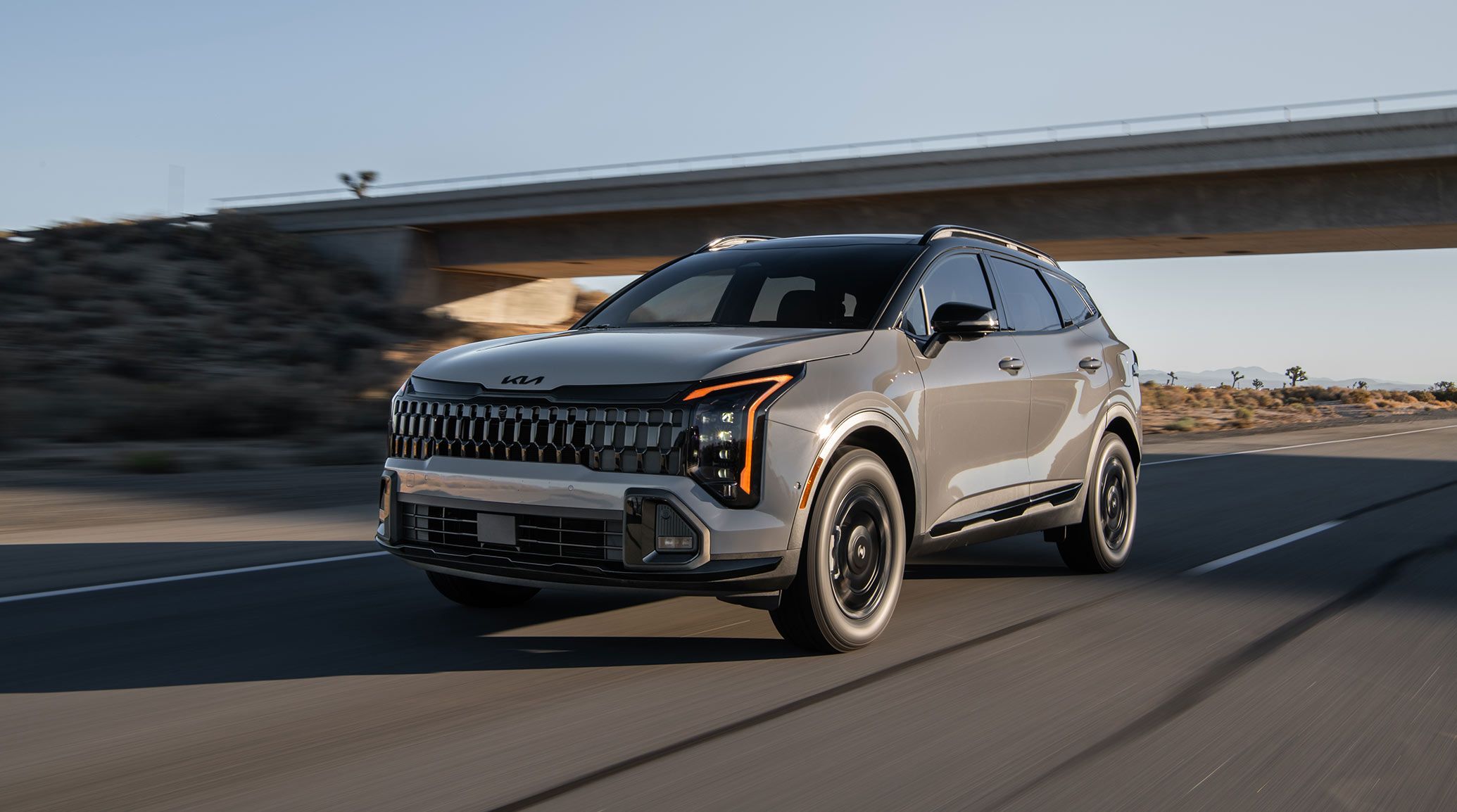 Photograph of the 2027 Kia Sportage SUV driving out from underneath a concrete bridge in the desert. The LED cube projector headlights are visible from the front corner.