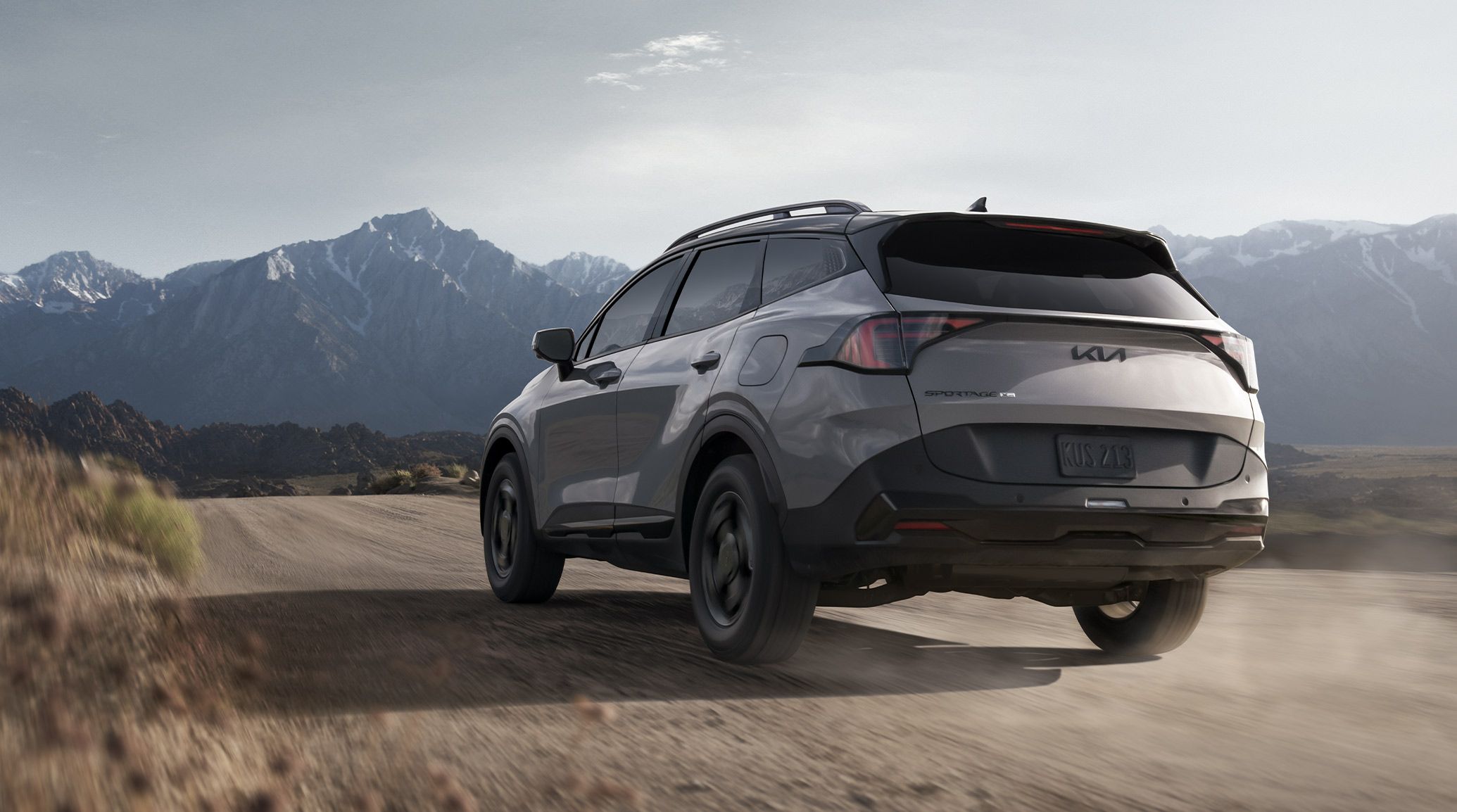 Rear-angled photograph of the 2027 Kia Sportage SUV in grey, traversing a gravel road in the desert with impressive, snow-lined mountains in the background. The rear spoiler and details are prominent on the back of the Sportage.