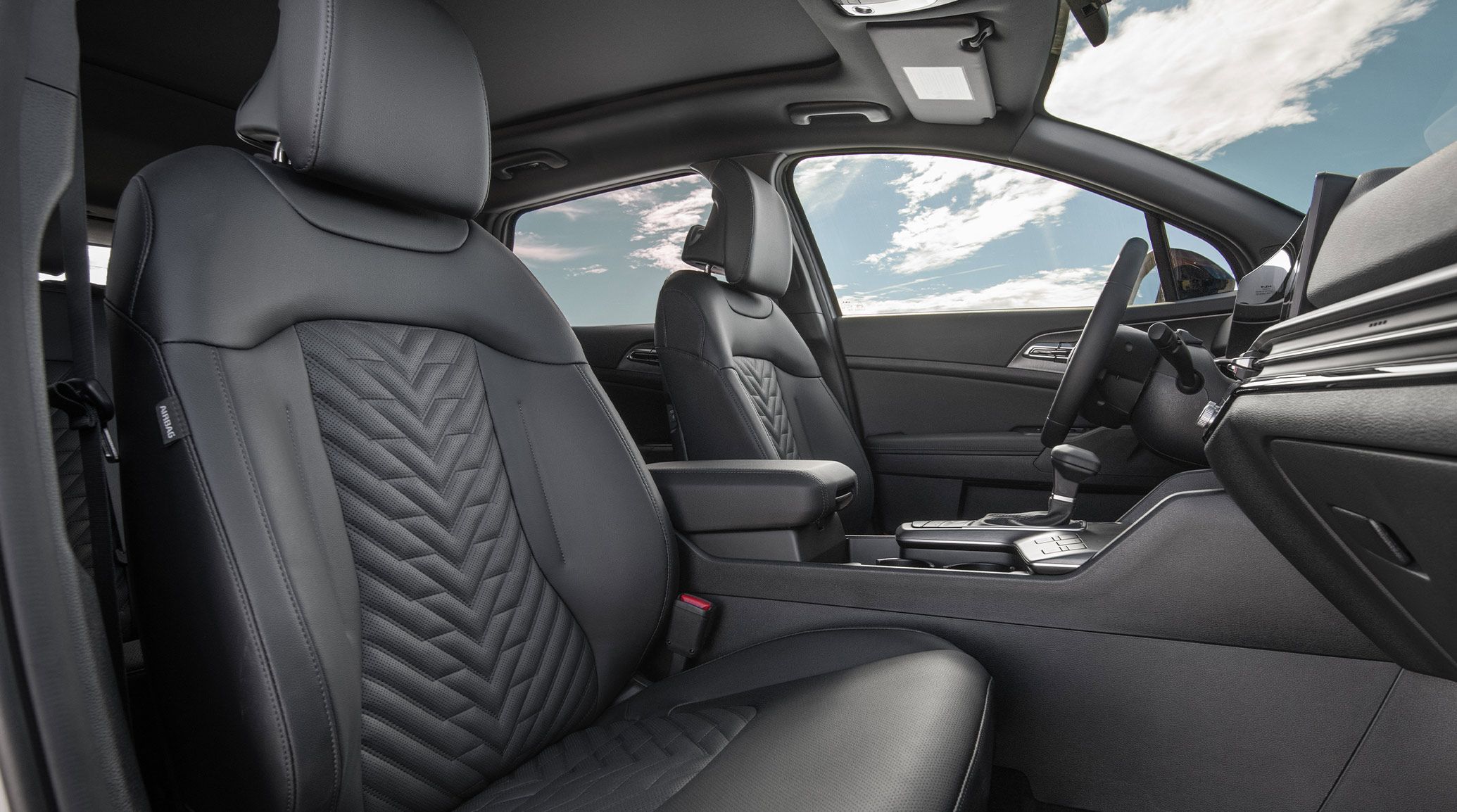 Interior view of the 2027 Kia Sportage SUV showcasing the perforated and quilted black heated and ventilated front seats. A blue sky with clouds and sun is visible through the windows.