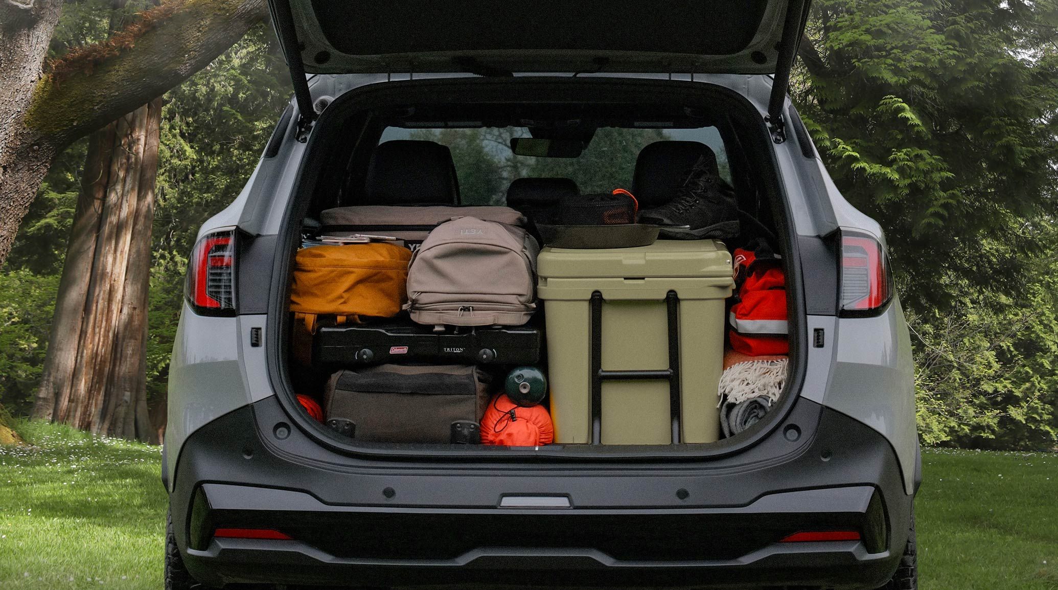 Rear shot of the 2027 Kia Sportage SUV with the trunk door opened upwards and the trunk filled with luggage, bags, totes, shoes, and other camping supplies, meant to highlight the impressive cargo room even with the 2nd-row seats up, headrests visible behind the bags. The SUV is parked in a grassy environment with lush trees in the background.