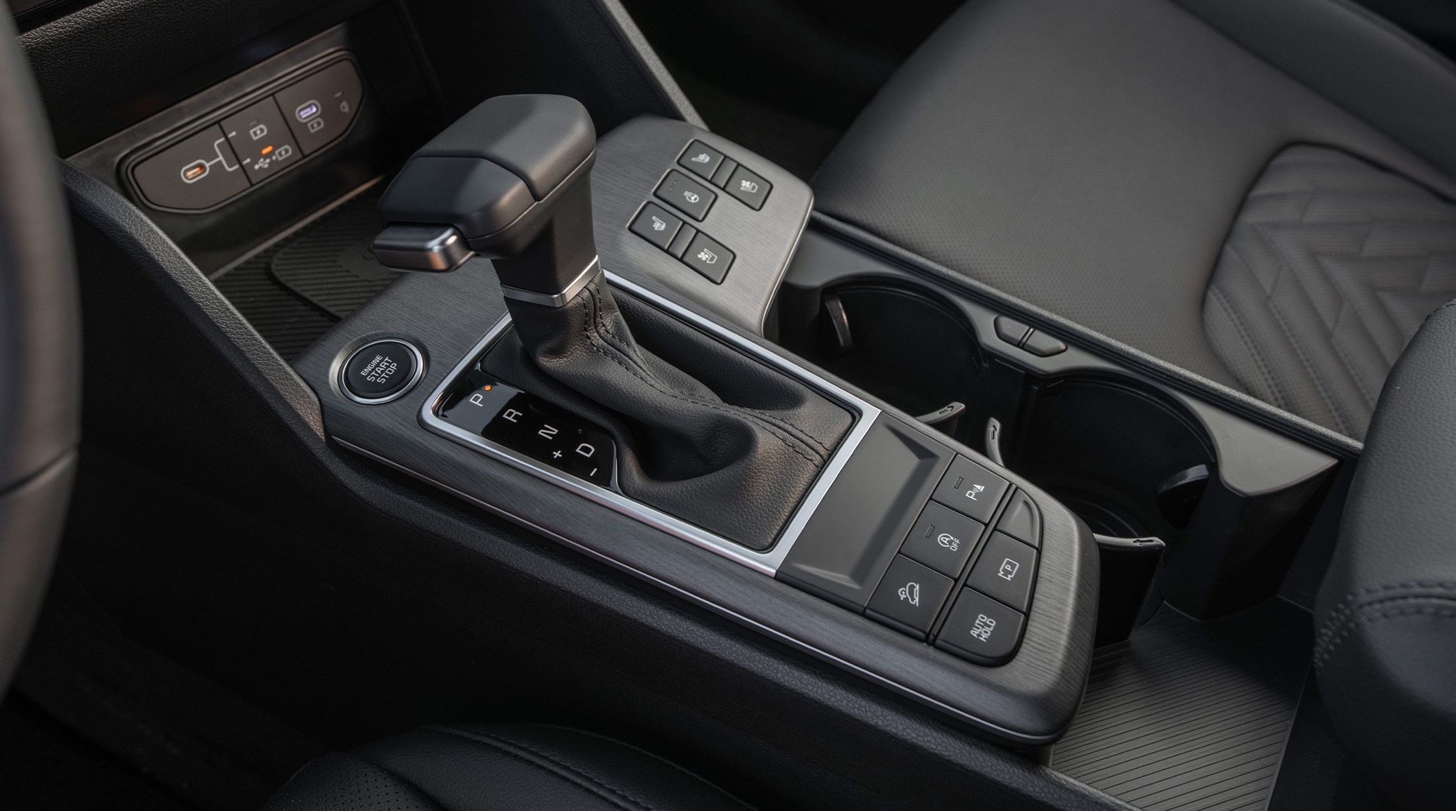 Close-up photograph inside the 2027 Kia Sportage SUV highlighting the automatic gear shifter and the related buttons that are nearby in the center area. Two large cupholders and the Engine Stop/Start button are also visible.
