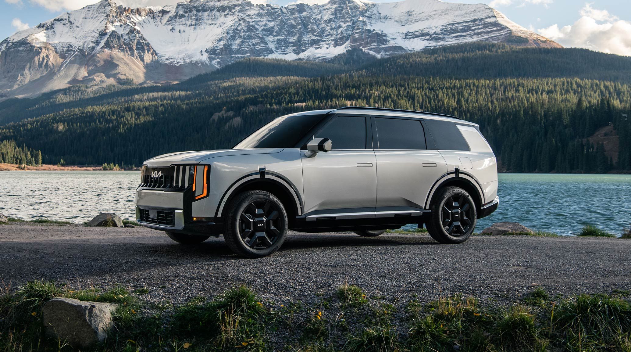 A still shot of the 2027 Kia Telluride Hybrid in silver parked on a rocky lakeside with towering snow-capped mountains and a dense forest in the background, highlighting the driver’s side with bold headlights and all-terrain tires.