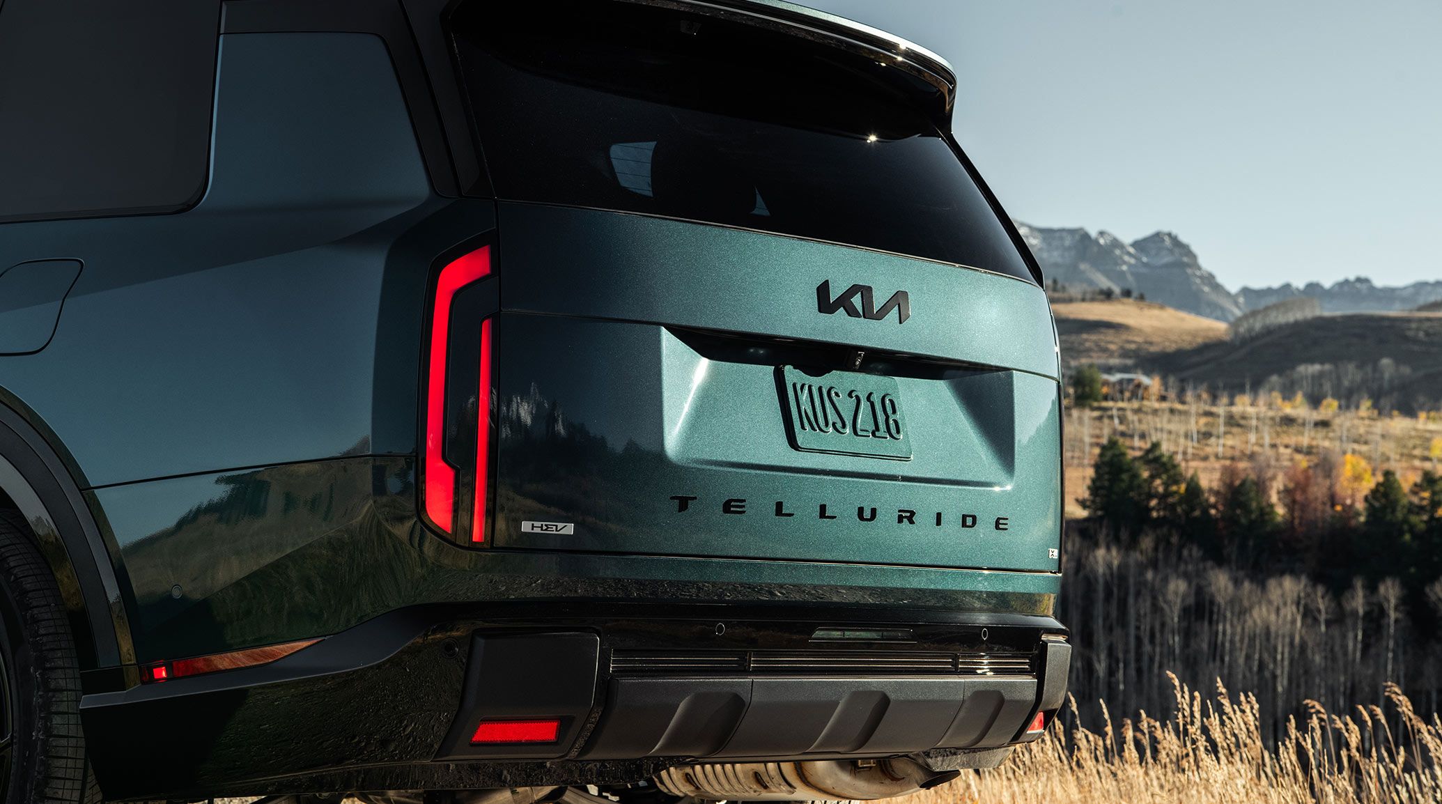 A close-up rear-view shot of the 2027 Kia Telluride Hybrid in blue-green, highlighting the brake lights and spacious trunk design. The SUV is against a grassy meadow.