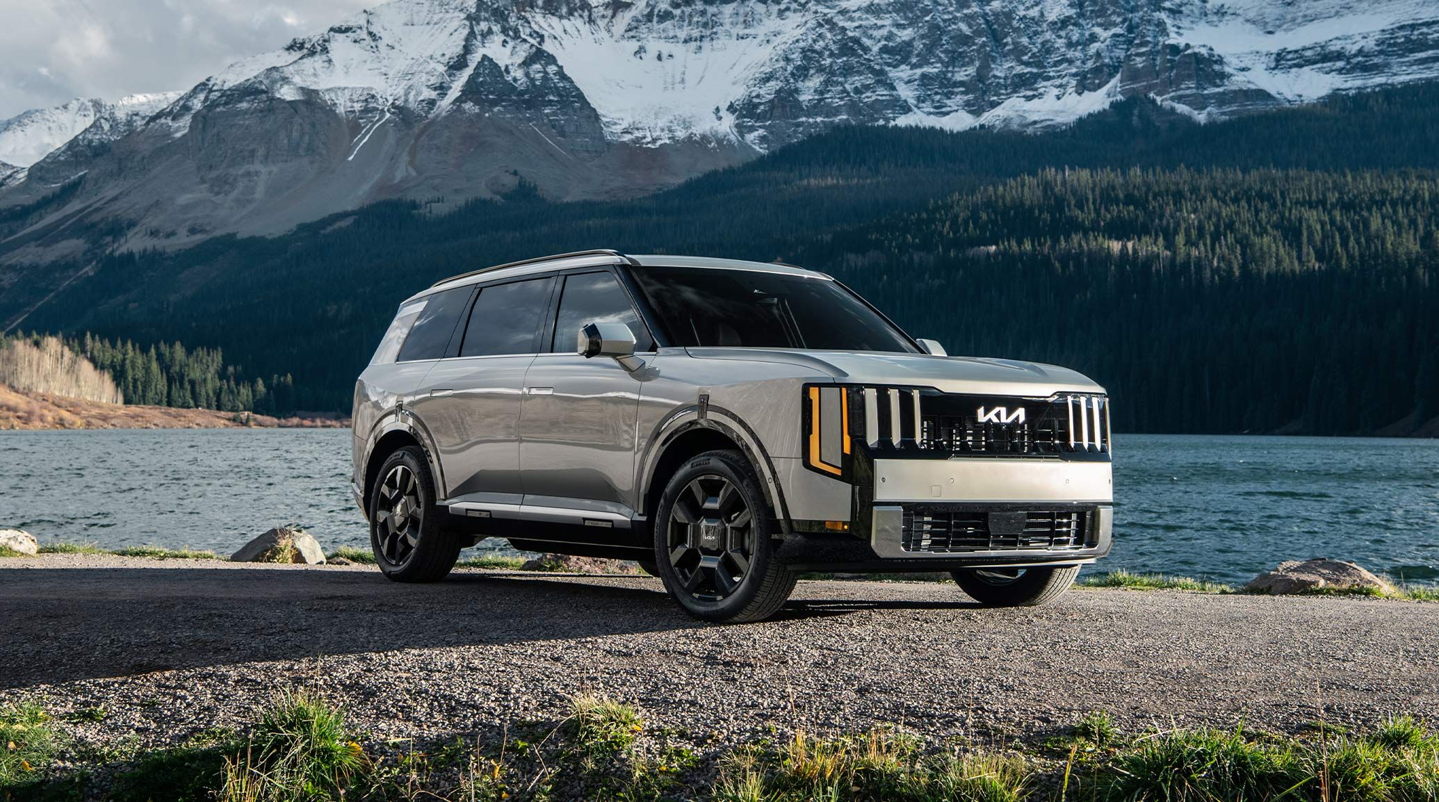 A silver 2027 Kia Telluride Hybrid parked on a rocky lakeside with towering snow-covered mountains in the background, highlighting the SUV’s bold exterior design and rugged capabilities.