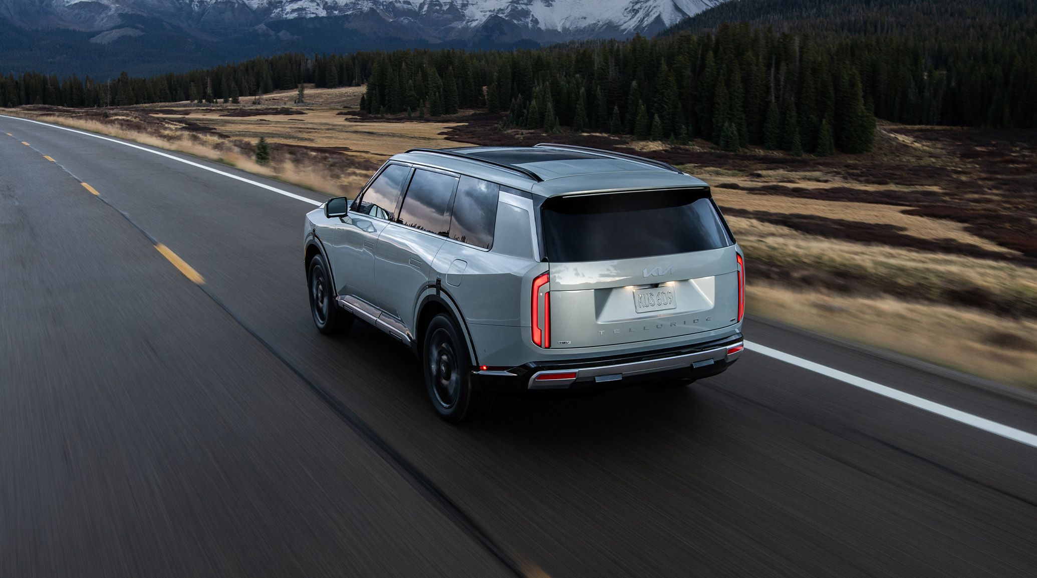 Rear three-quarter exterior view of the 2027 Kia Telluride Hybrid in light silver driving on an open highway, with LED vertical taillights, low-profile roof rails, and a rugged SUV design set against a scenic mountain landscape.