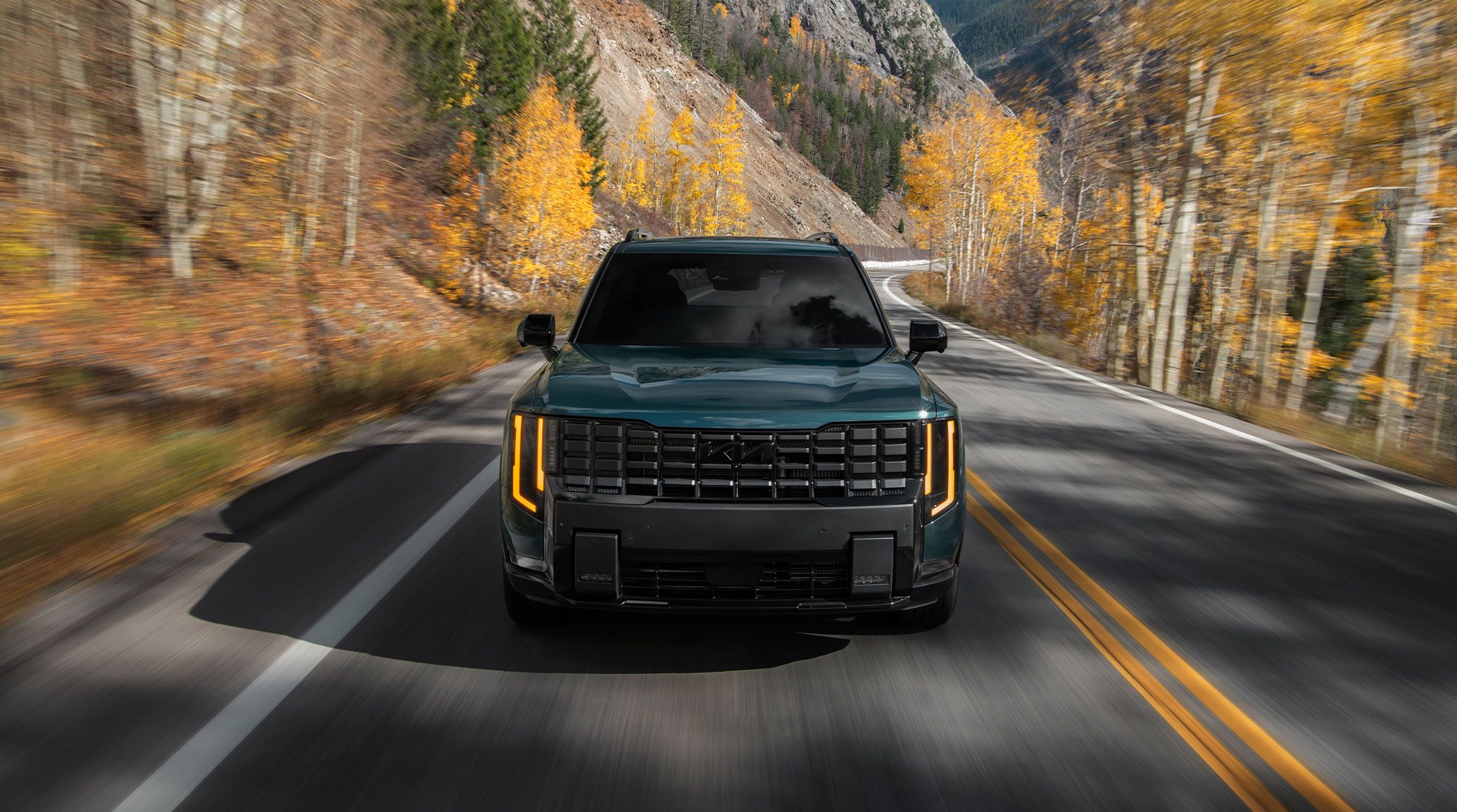 A front-facing view of the 2027 Kia Telluride Hybrid in blue-green driving on a highway, framed by autumn trees in the background. The image highlight's the SUV's tinted front windshield and bold black grille.