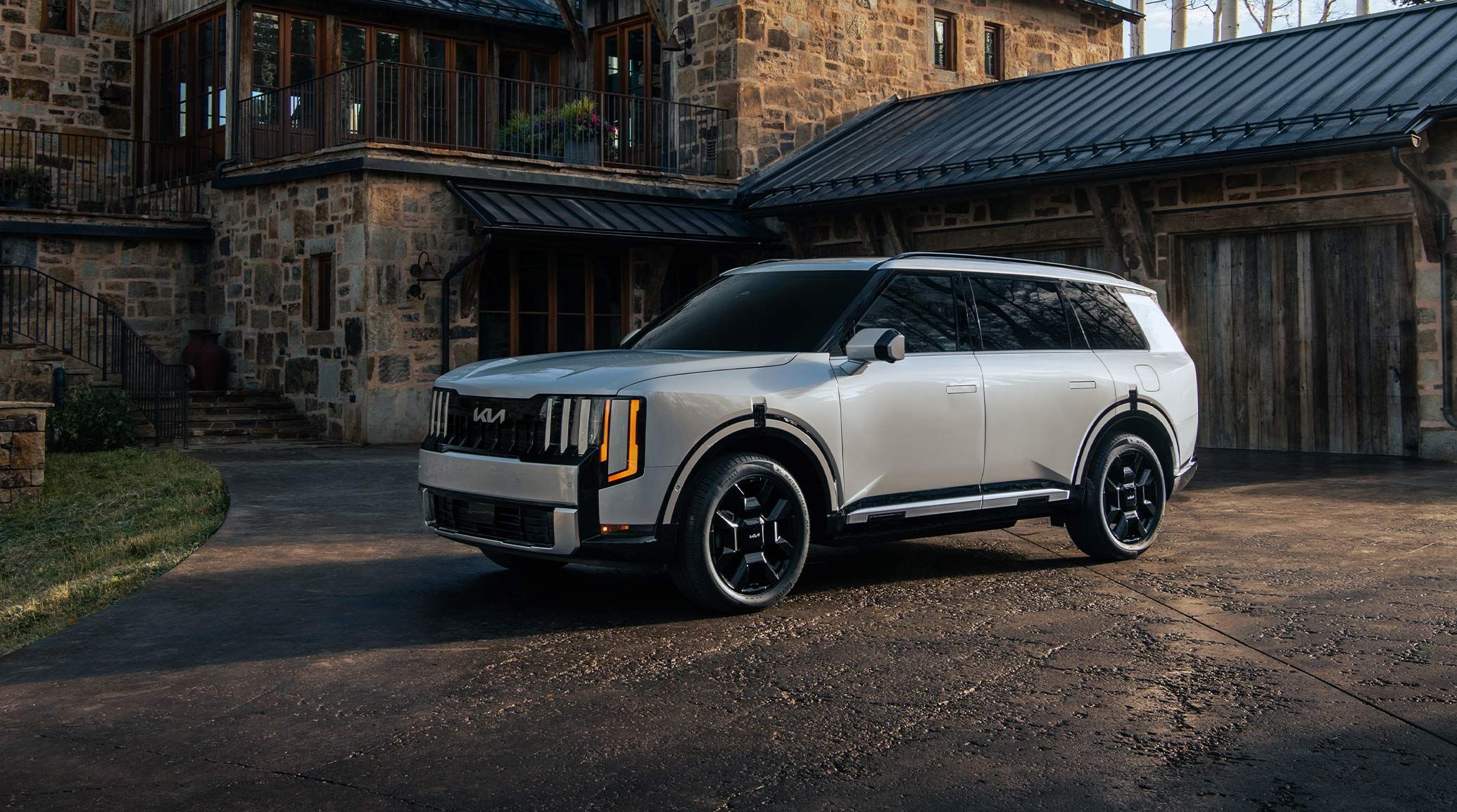 Front three-quarter exterior view of the 2027 Kia Telluride Hybrid in silver parked in a rustic driveway, with a bold grille, black alloy wheels, and auto-extending handles.
