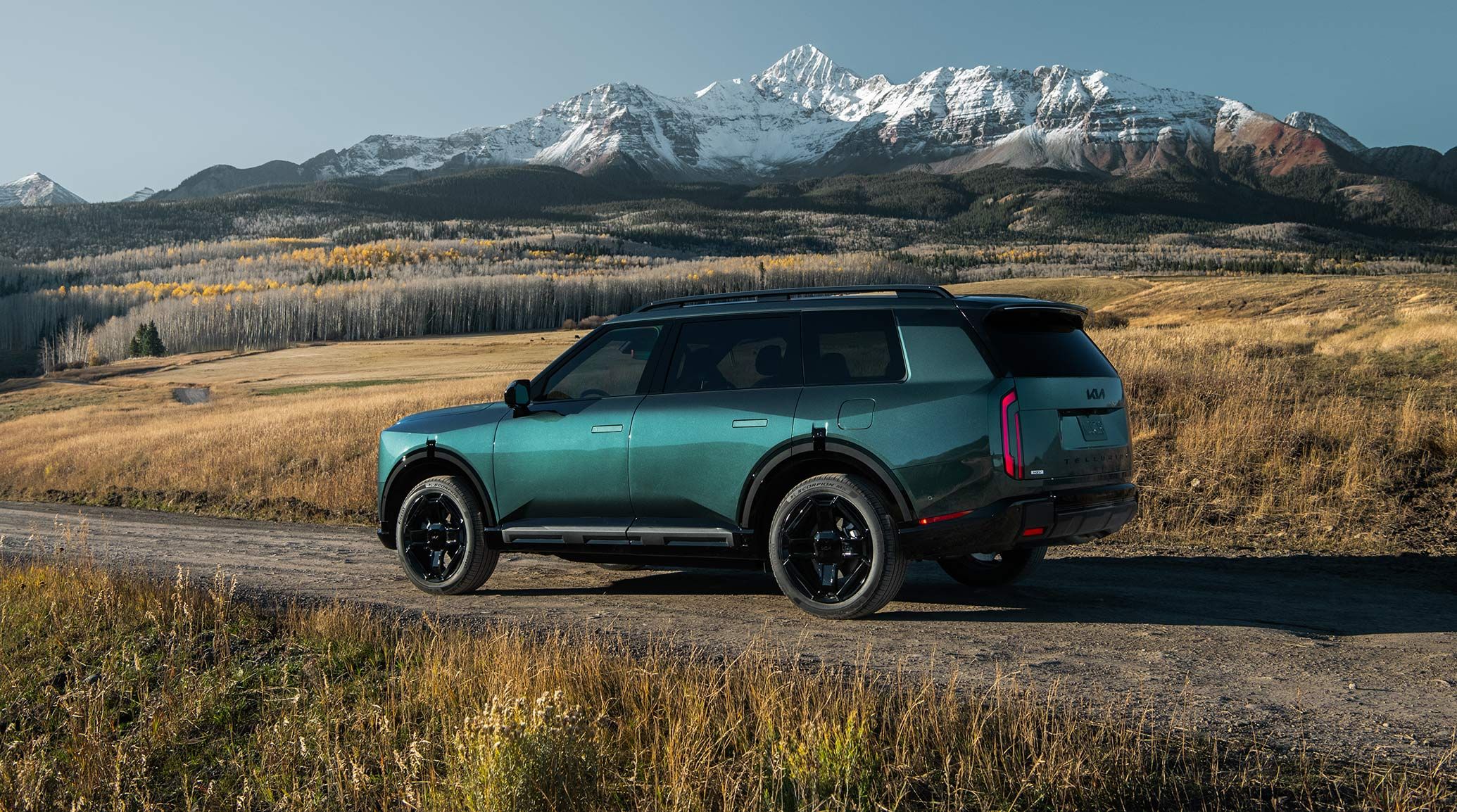 The 2027 Kia Telluride Hybrid SUV is shown in blue-green on a dirt road with rugged terrain and snowy mountains behind. The driver-side is facing front, highlighting the rear design for spacious cargo and tinted windows for privacy.