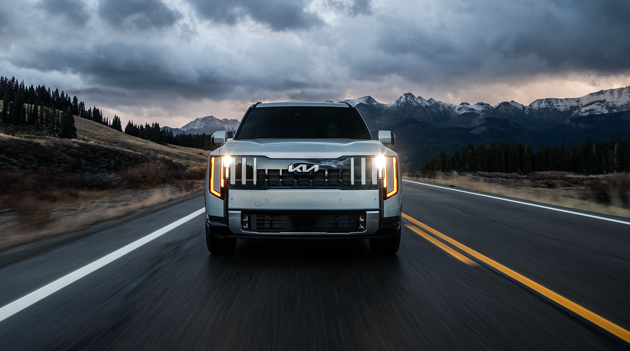 An action shot of the 2027 Kia Telluride Hybrid in silver driving along a highway with mountains, dense trees, and cloudy skies behind it. The front of the SUV is highlighted with LED headlights on.