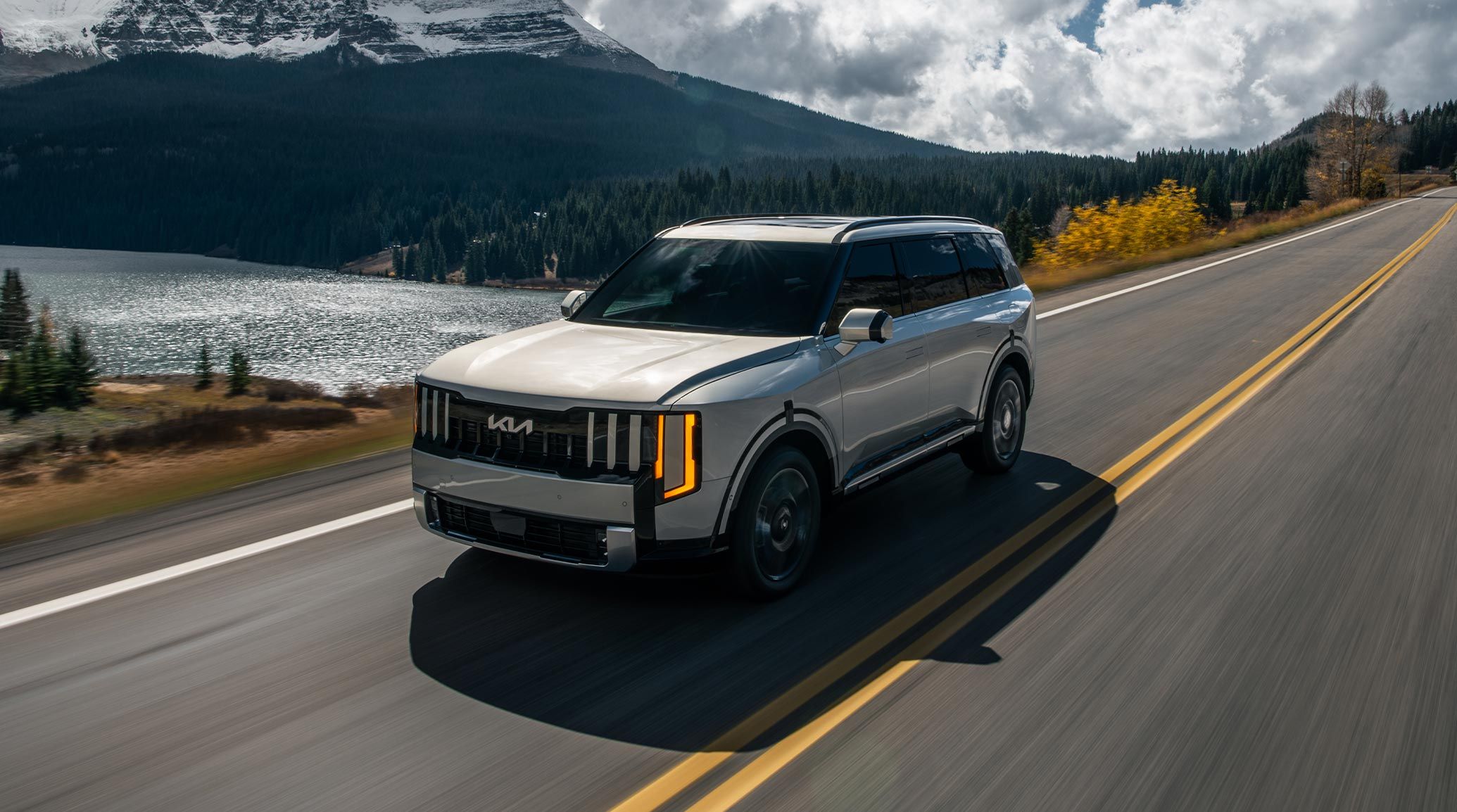A shot of a silver 2027 Kia Telluride Hybrid driving along a highway with a scenic lakeside view and mountains beside it. The front of the SUV is highlighted, showcasing its rugged design.