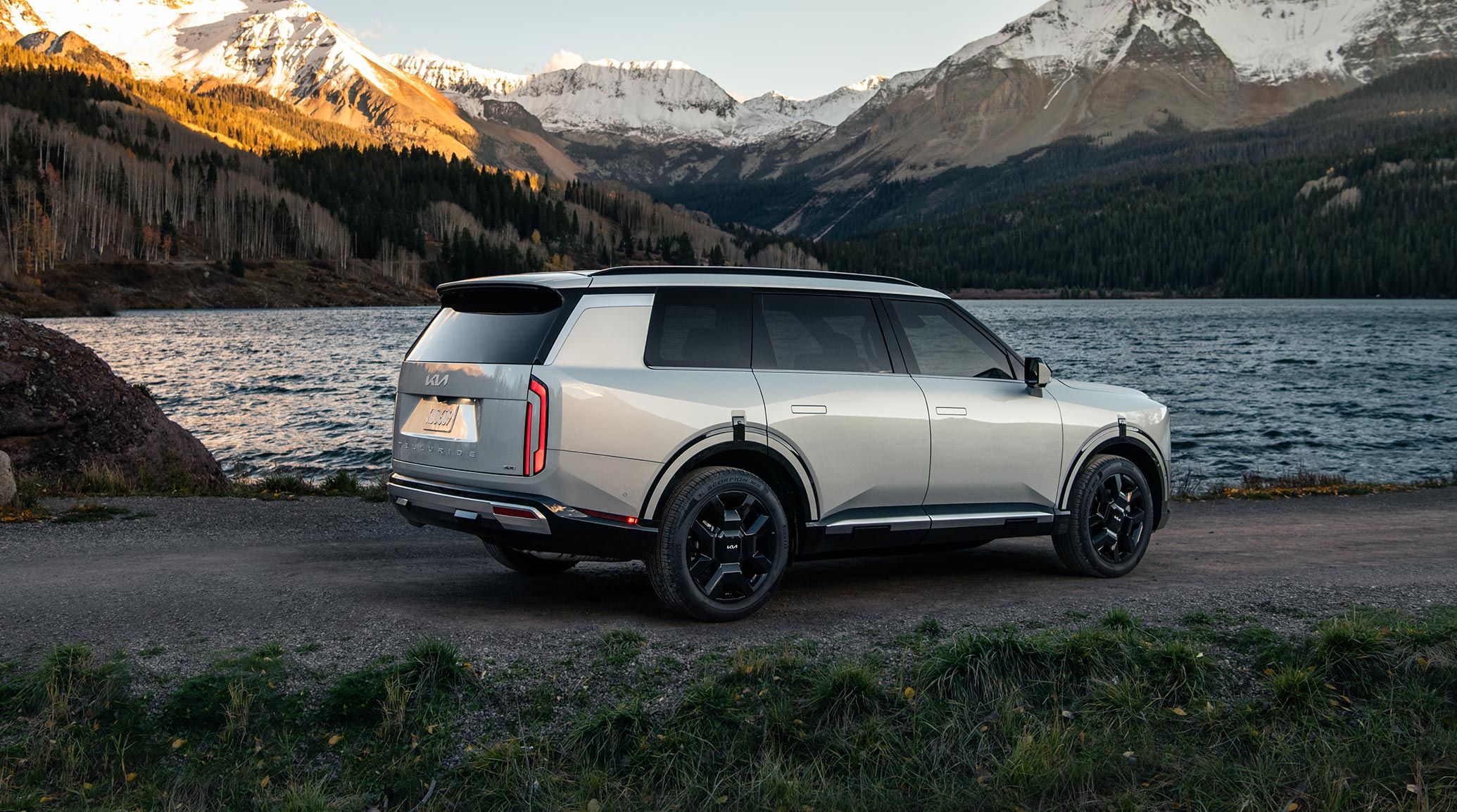 A rear-side view of the 2027 Kia Telluride Hybrid SX Prestige in silver parked on a dirt path next to a scenic lakeside with snowy mountains behind it, showcasing its tinted windows, alloy wheels, and off-road abilities.