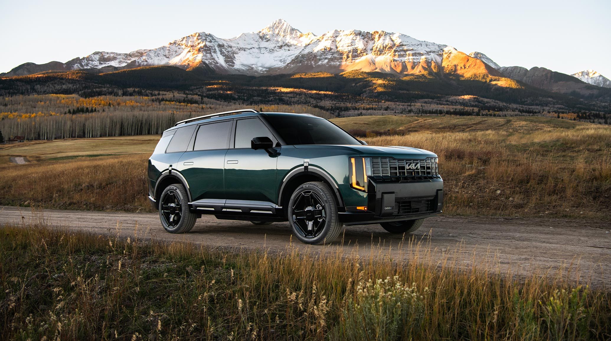 A still image of the 2027 Kia Telluride Hybrid in a jade green exterior, positioned on a dirt path with the passenger side facing the camera. The SUV is surrounded by plants and set against snowy mountain peaks.