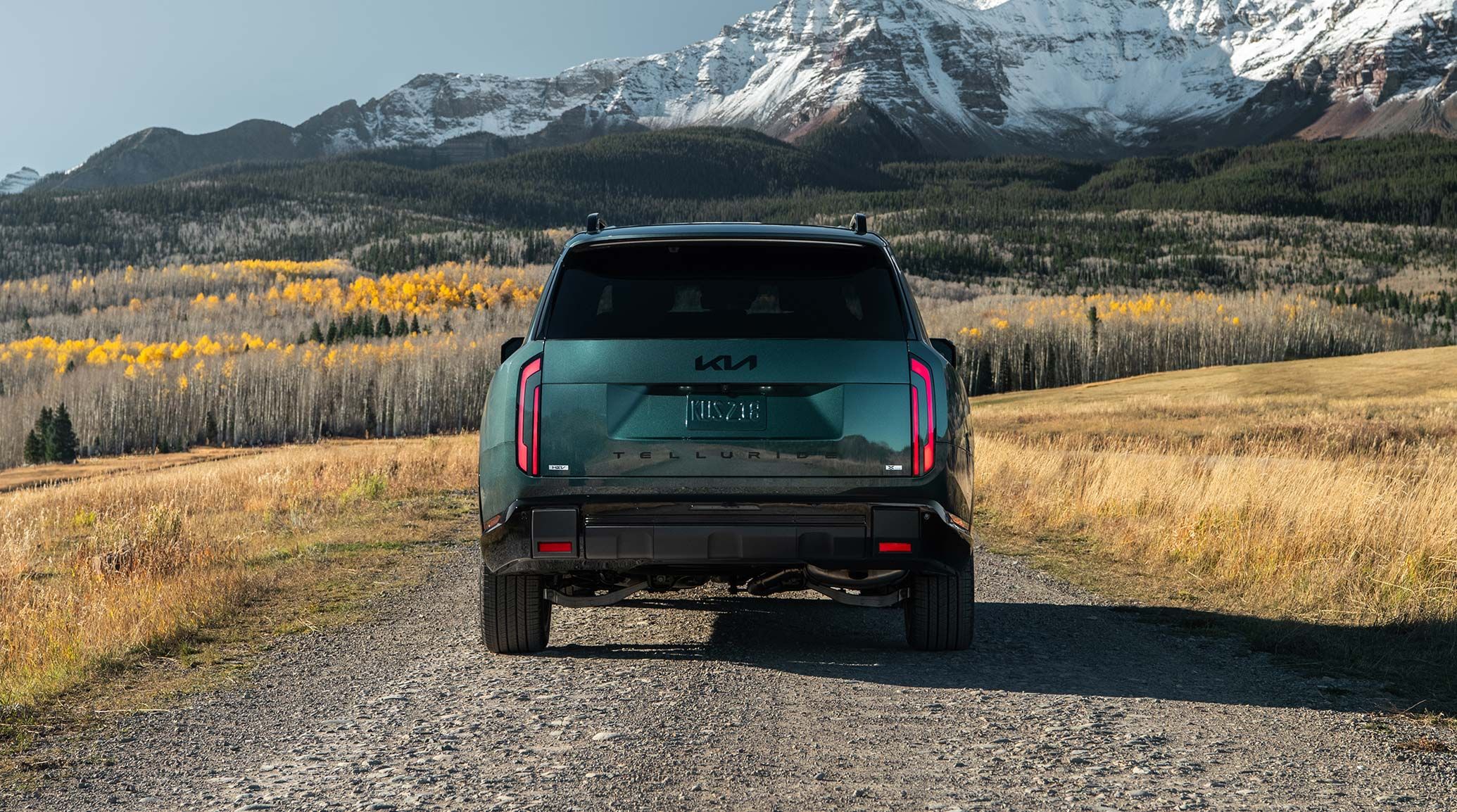 A rear-view shot of the 2027 Kia Telluride Hybrid X-Line SX Prestige in jade green stopped on a dirt path, with brake lights on and a tinted rear window. Plants and snowy mountains fill the background.
