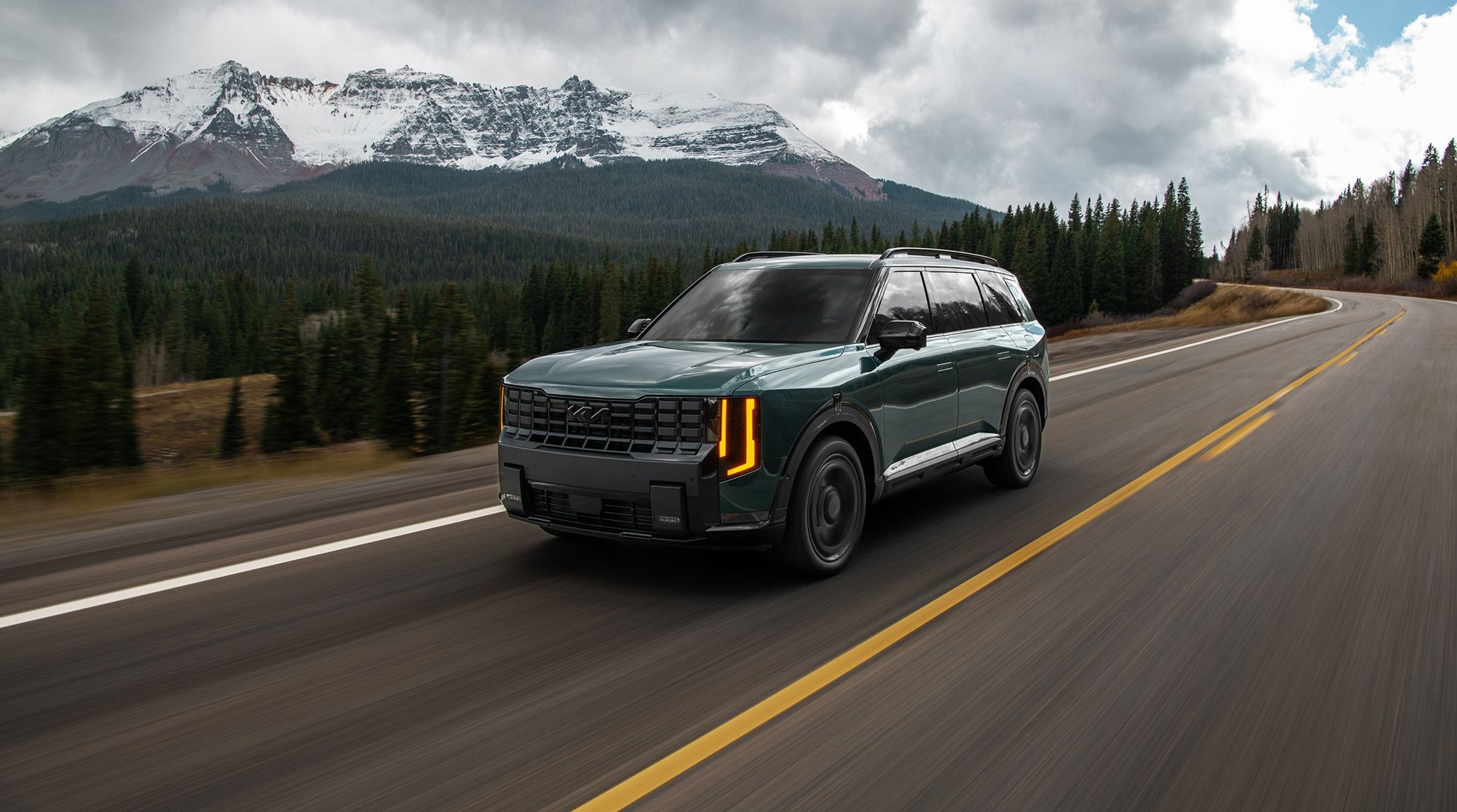The 2027 Kia Telluride Hybrid in jade green driving on a highway with snowy mountains and lush trees behind. The driver-side is facing out, highlighting the bold grille, LED headlights, and tinted windows for privacy.
