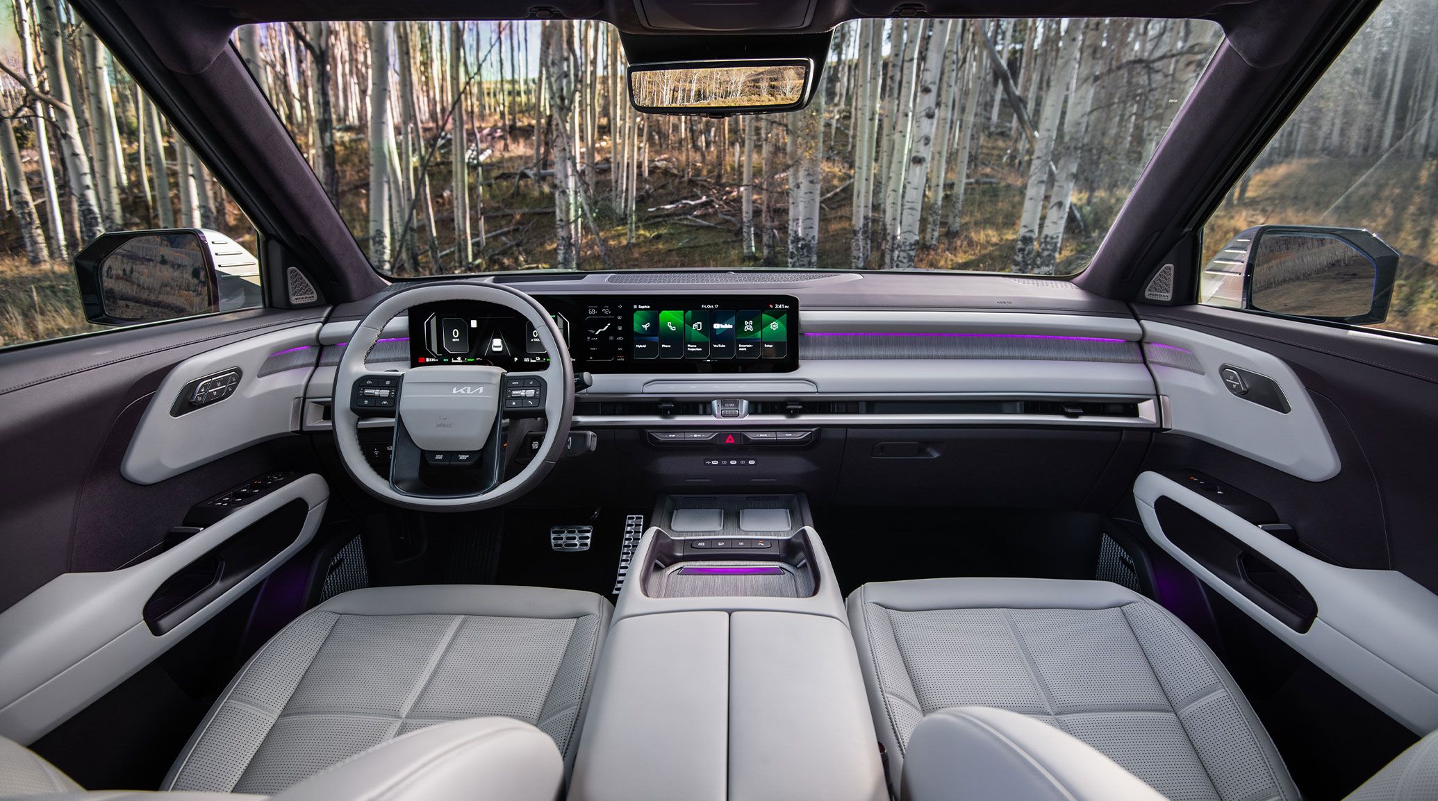 An interior view of the 2027 Kia Telluride Hybrid in sand beige, highlighting the front suede seat trim, steering wheel, digital display, and cup holders with trees visible outside.