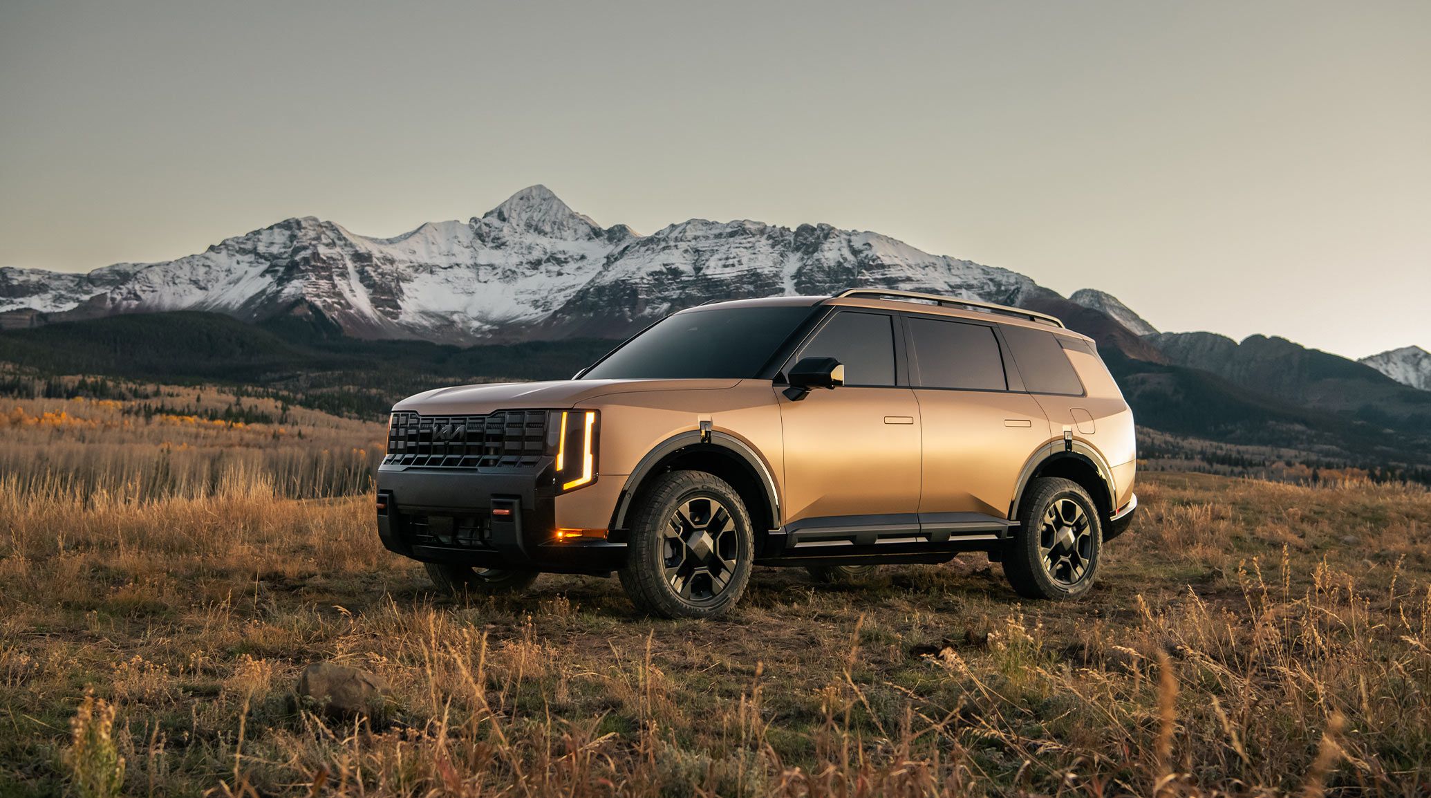 A side view of the 2027 Kia Telluride X-Pro in brown parked in a grassy field with snowy mountains in the background.