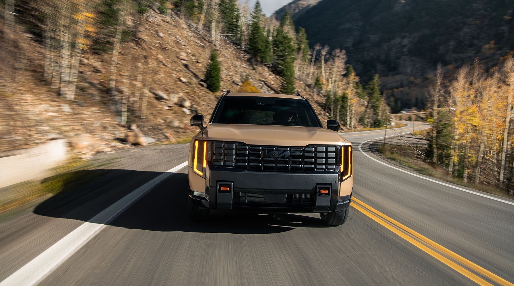 Dynamic action shot of the 2027 Kia Telluride in terrain brown speeding along a winding mountain road, showcasing the bold front grille, sleek side mirrors, and powerful performance.