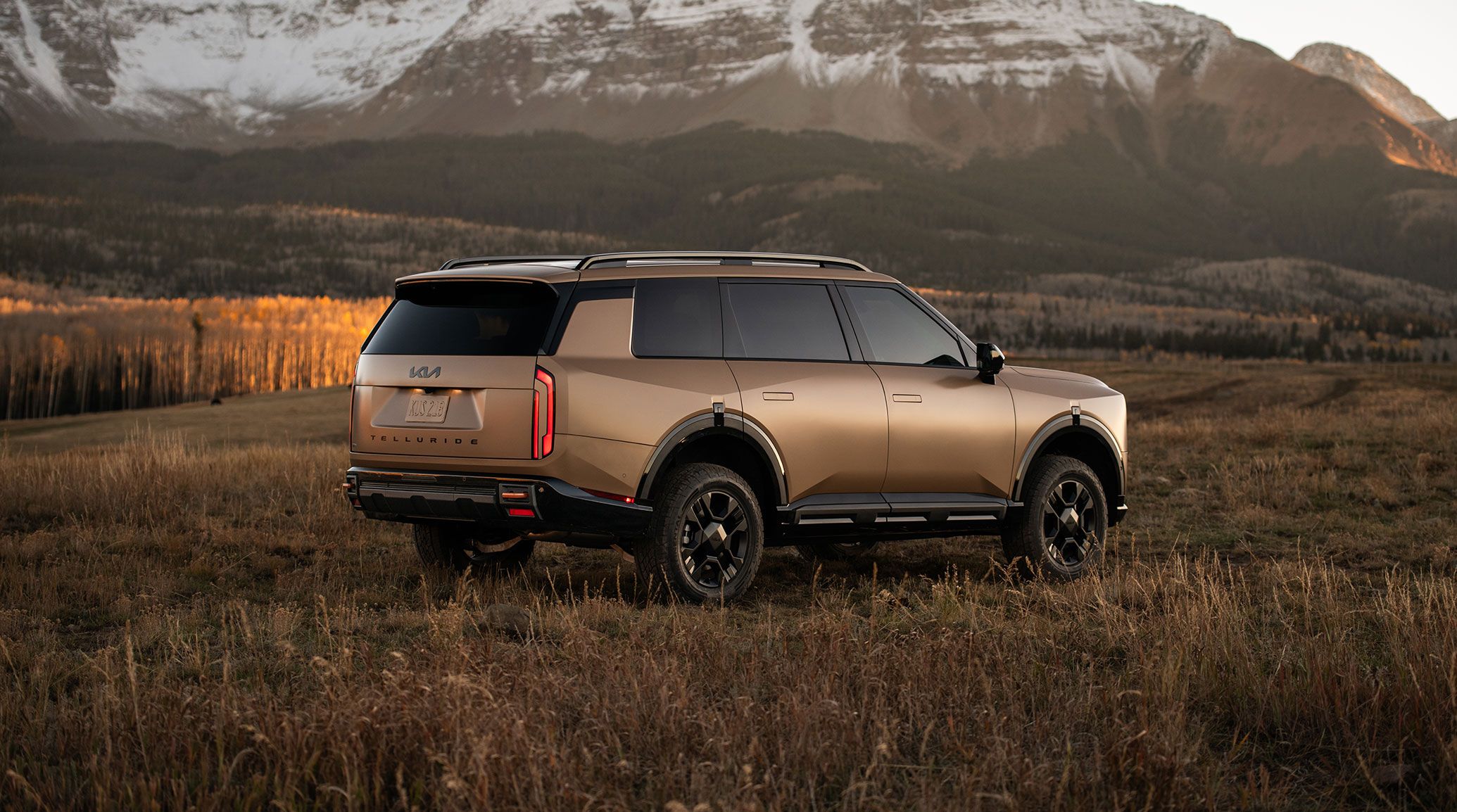 Passenger-side and rear view of the 2027 Kia Telluride in brown, showcasing stylish alloy wheels, sleek LED taillights, and rugged SUV design, parked on a grassy field with snow-covered mountains in the background.