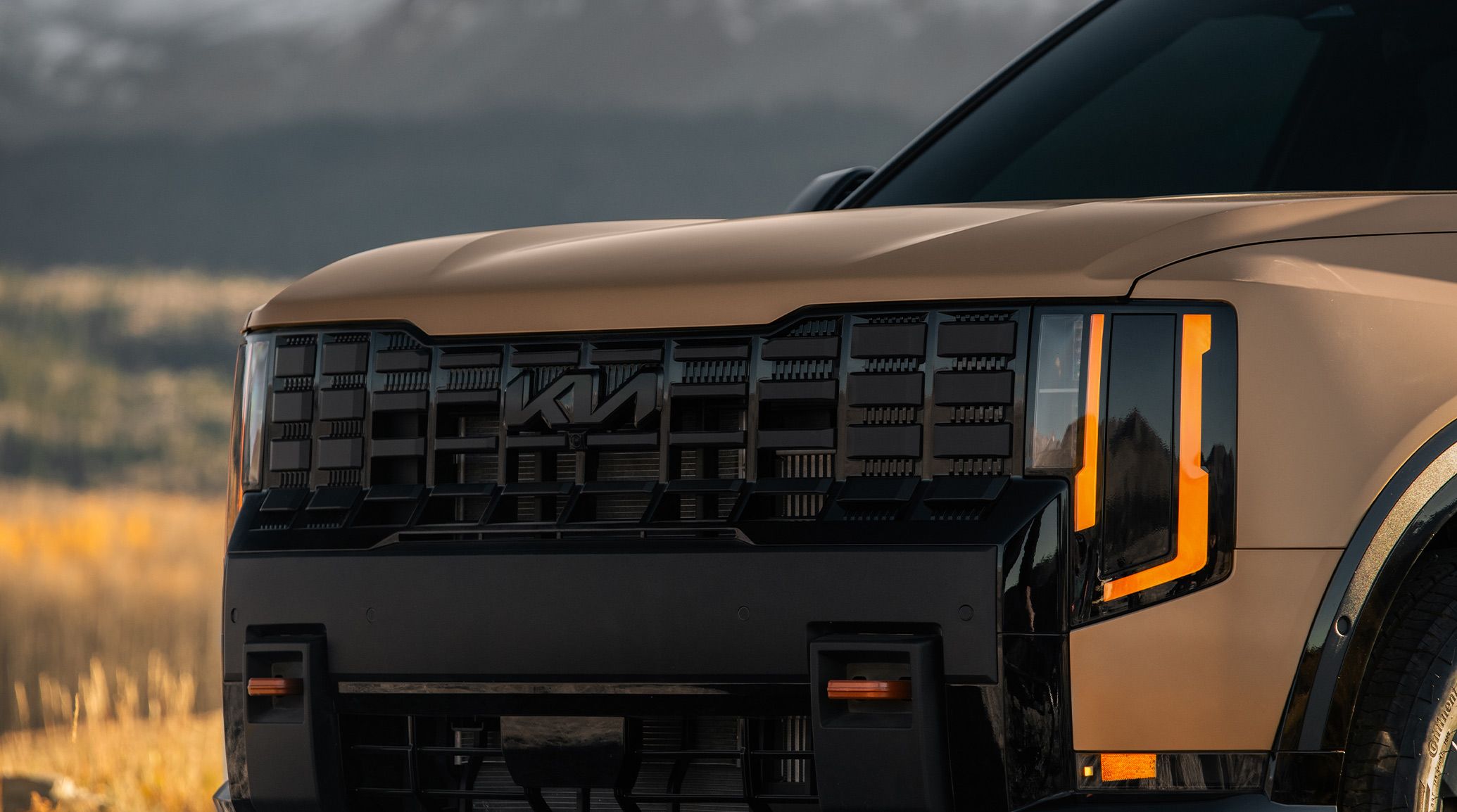 Close-up shot of the 2027 Kia Telluride in brown featuring a bold black front grille and dual front recovery hooks, set against a backdrop of vibrant fall foliage.