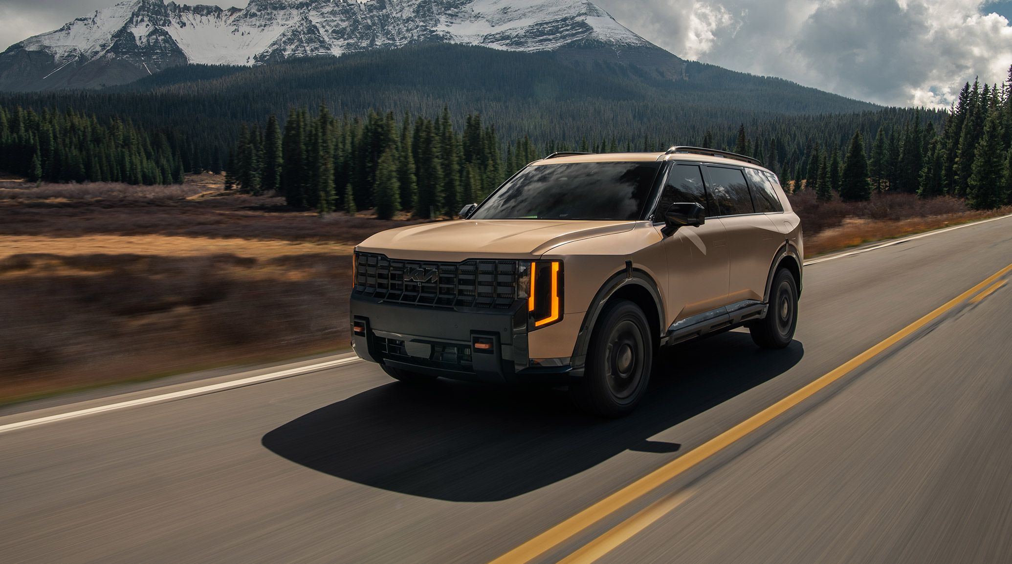 Angled shot of the 2027 Kia Telluride X-Pro SX Prestige in brown, highlighting gloss black accents and rugged matte wheel arches while cruising through a forested mountain road.