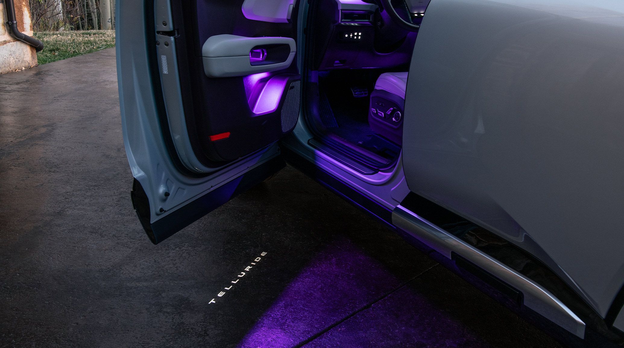 An image of the 2027 Kia Telluride in gray parked in a driveway, highlighting the driver-side door with purple LED accent lights and illuminated Telluride logo projected onto the ground.