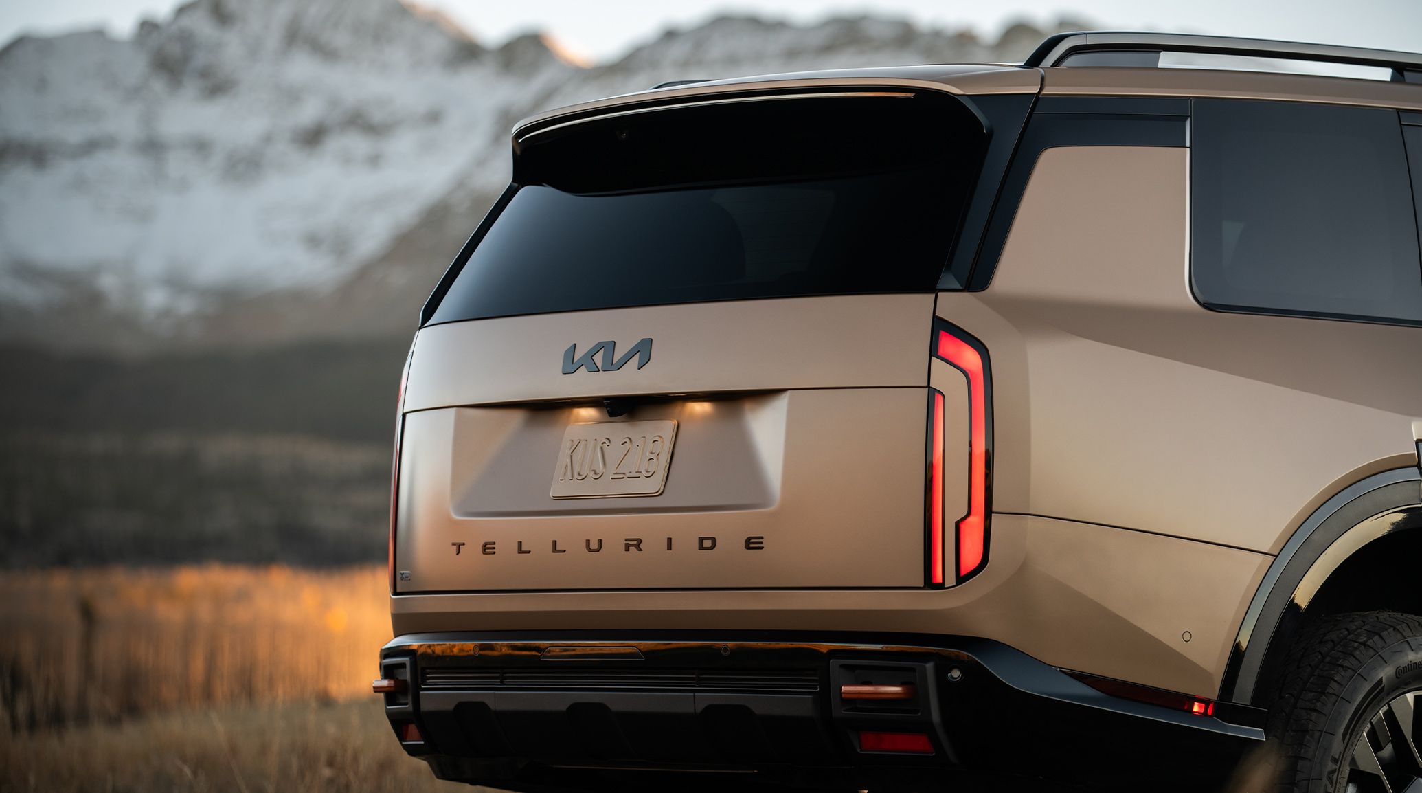 A still shot of the 2027 Kia Telluride in matte brown parked with the trunk closed, showcasing the SUV's sleek LED taillights against a backdrop of snow-covered mountains.