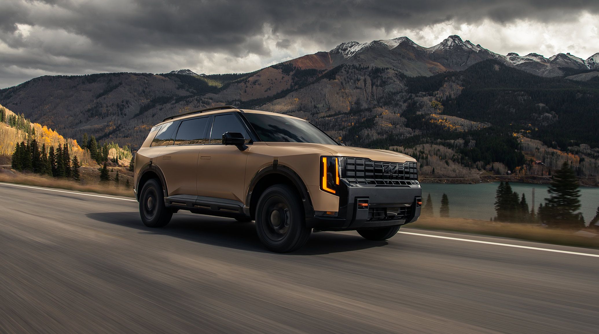 Dynamic shot of the 2027 Kia Telluride X-Pro SX Prestige in terrain brown speeding along a scenic mountain road with a lake in the background, featuring a bold front grille, striking vertical LED headlights, and rugged off-road styling.