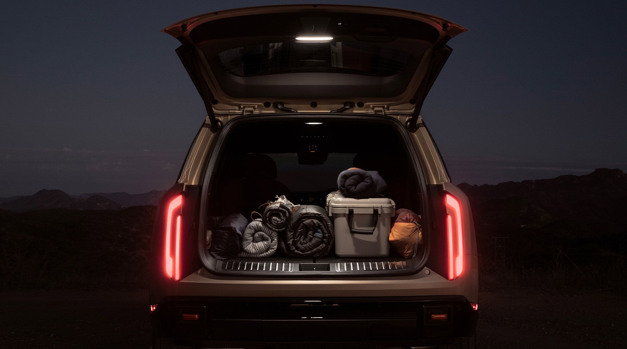 Photo of the 2027 Kia Telluride with an open trunk showcasing the spacious cargo area packed with outdoor gear, illuminated taillights, and liftgate lamp, set against a hillside backdrop.