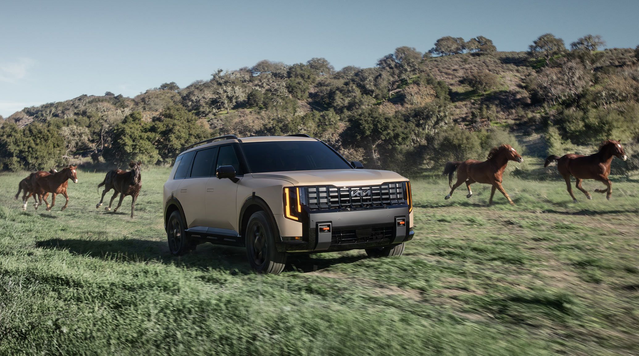 2027 Kia Telluride X-Pro SX Prestige in brown driving on grass with hills and bushes behind it. There are four horses running alongside the SUV, and the shot highlights the black windows and diverse terrain that the vehicle can adapt to.