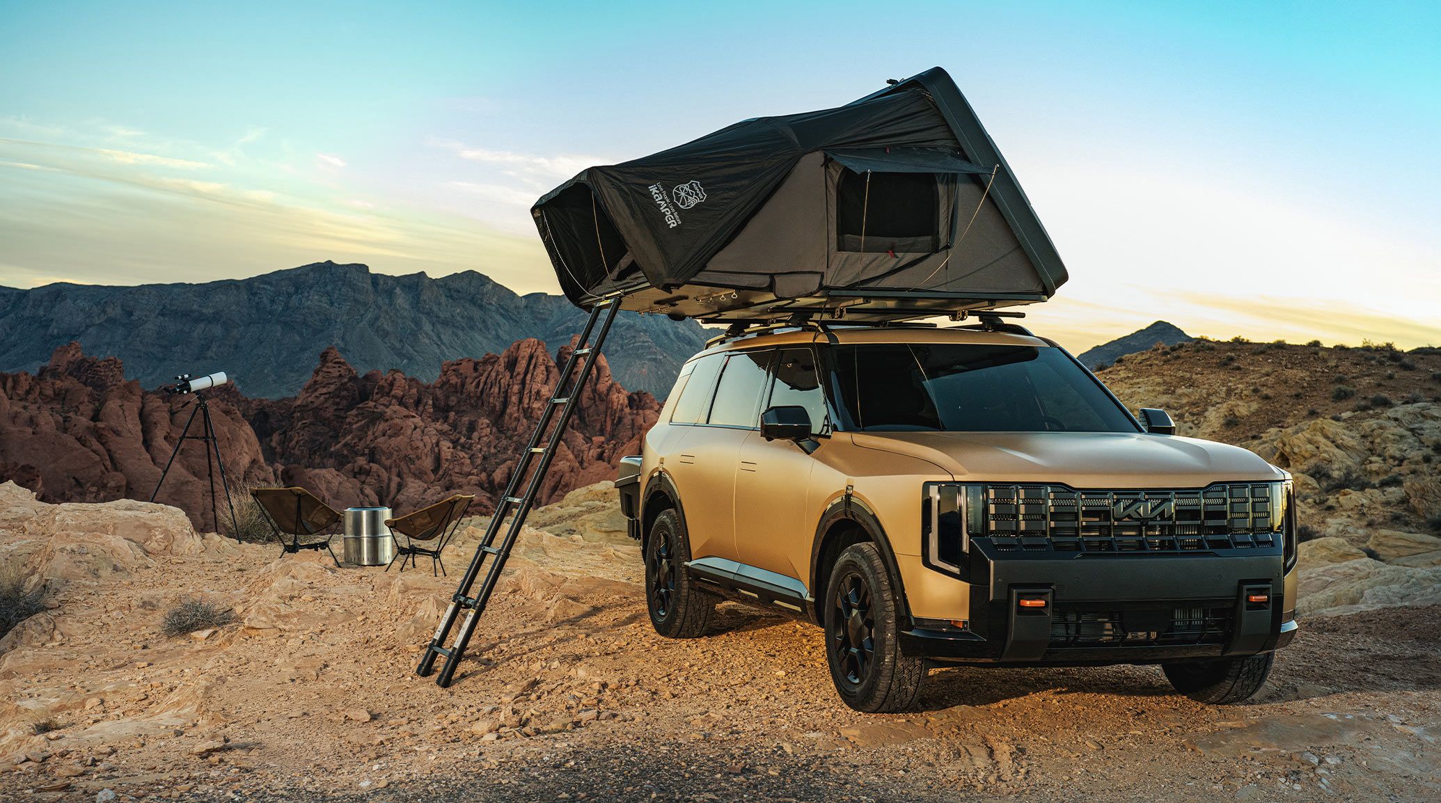 A 2027 Kia Telluride in brown is parked on rocky terrain with mountains in the background. A rooftop IKamper Skycamp tent is set up on cross bars, with a ladder extending to the ground. Two folding chairs and a telescope sit beside the SUV, creating a rugged campsite scene.