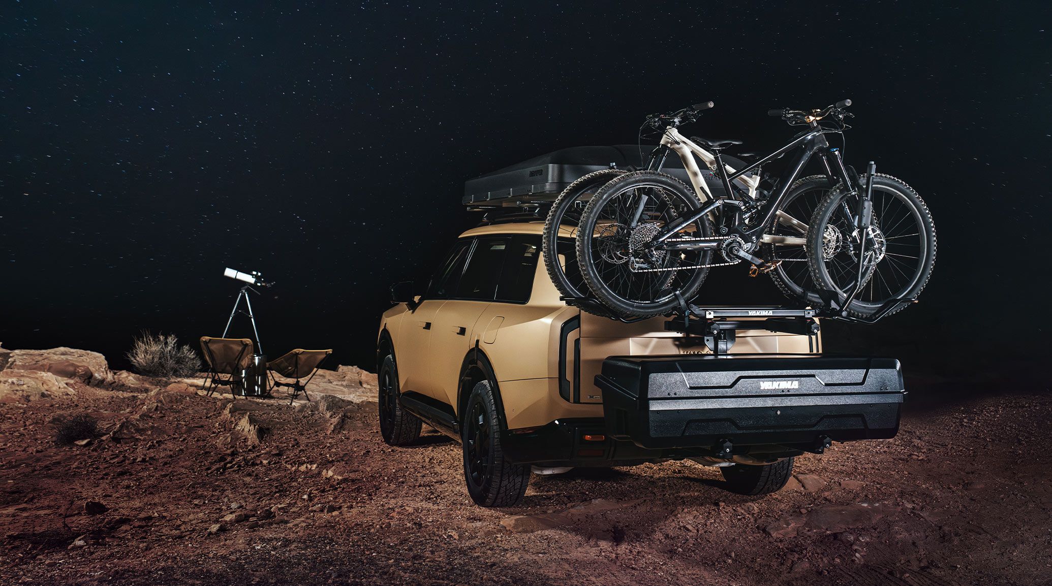 A still shot of the 2027 Kia Telluride rear with accessory cross bars, IKamper Skycamp, and a tow hitch bike attachment shown with 2 bikes on the vehicle. The SUV is parked on a cliff next to 2 folding chairs and a telescope with dark skies and stars visible.